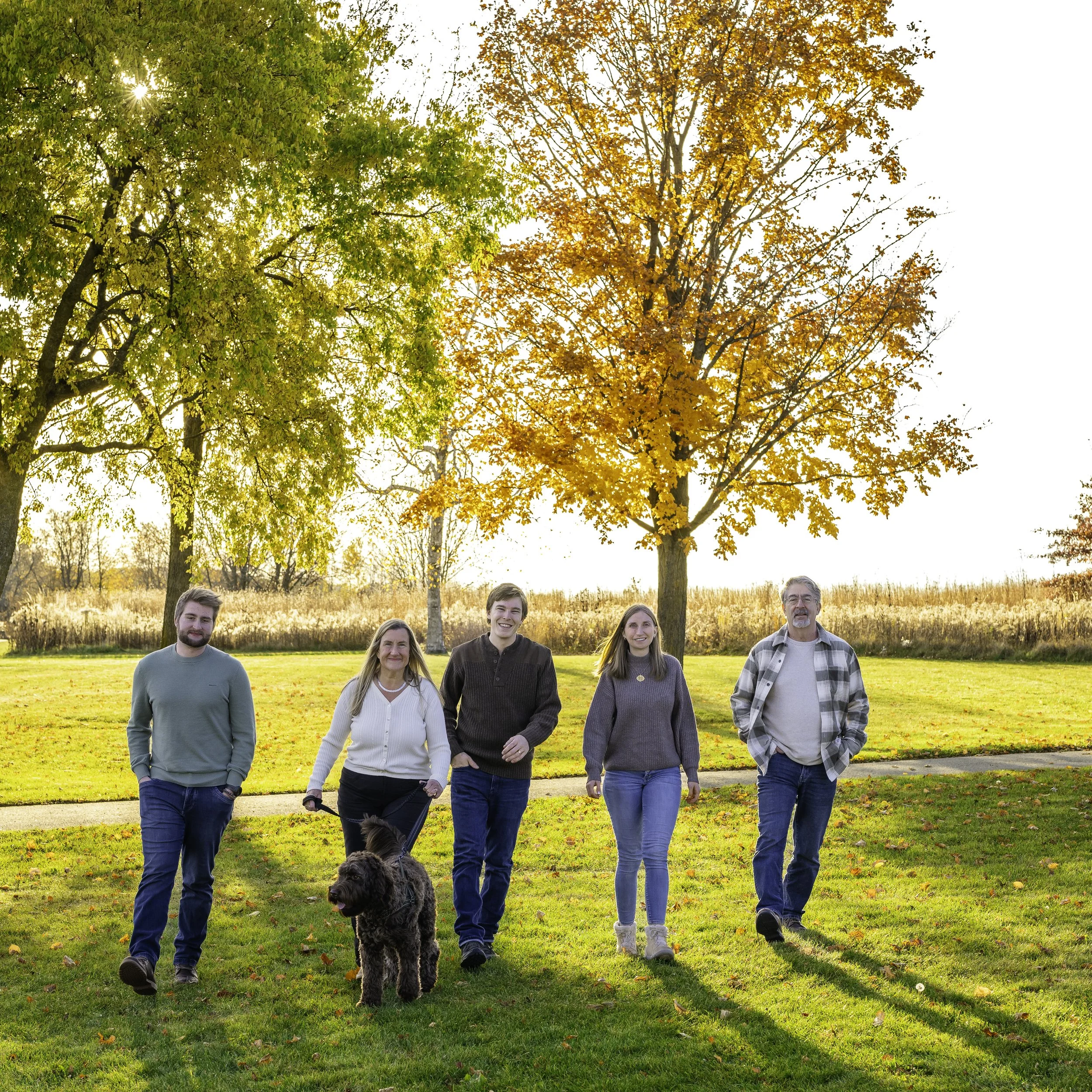 A group of five people and a dog walking in a park during fall, with colorful trees and a grassy area.