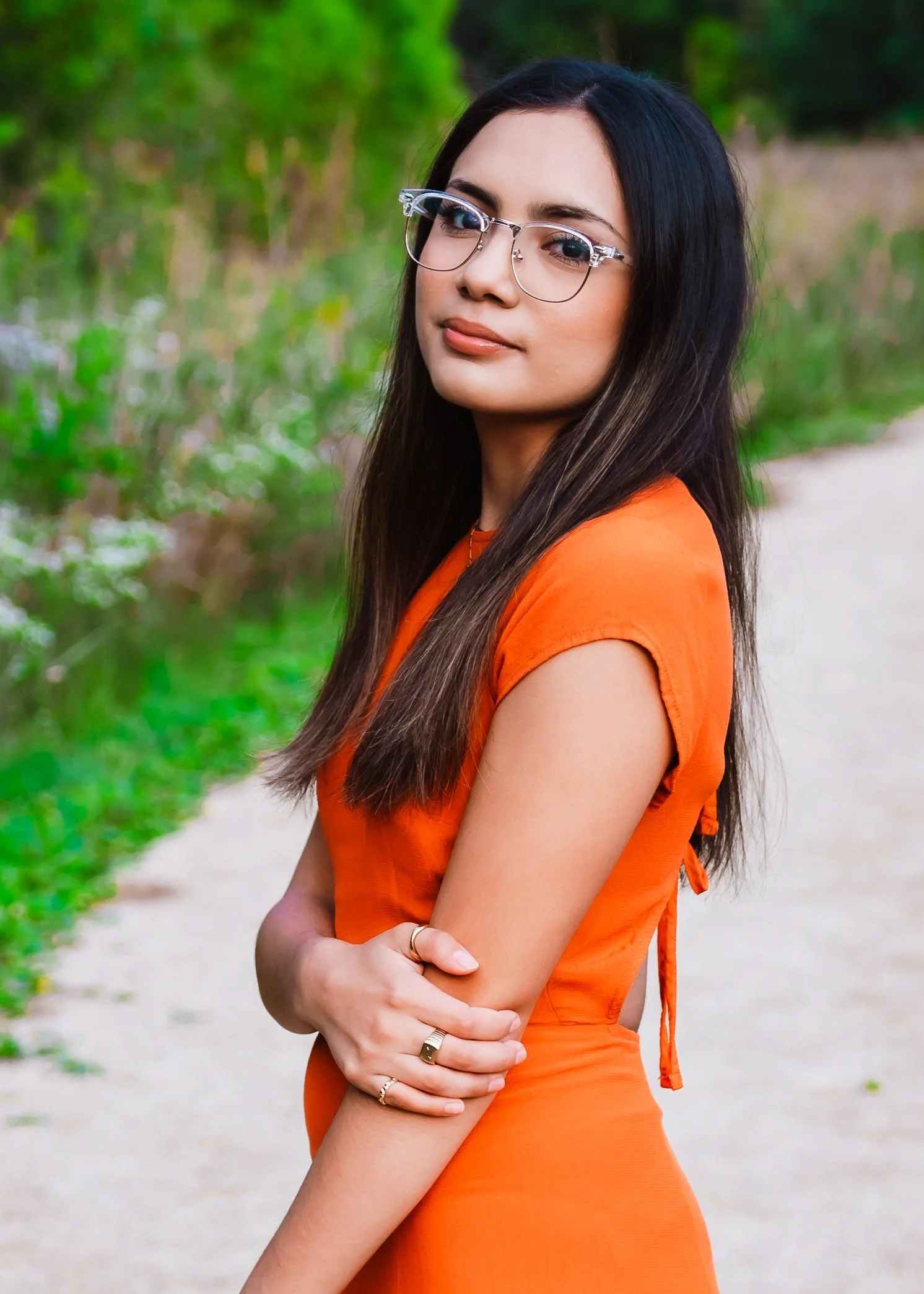 A young woman with long dark hair wearing glasses and an orange dress standing outdoors on a dirt path with greenery in the background.