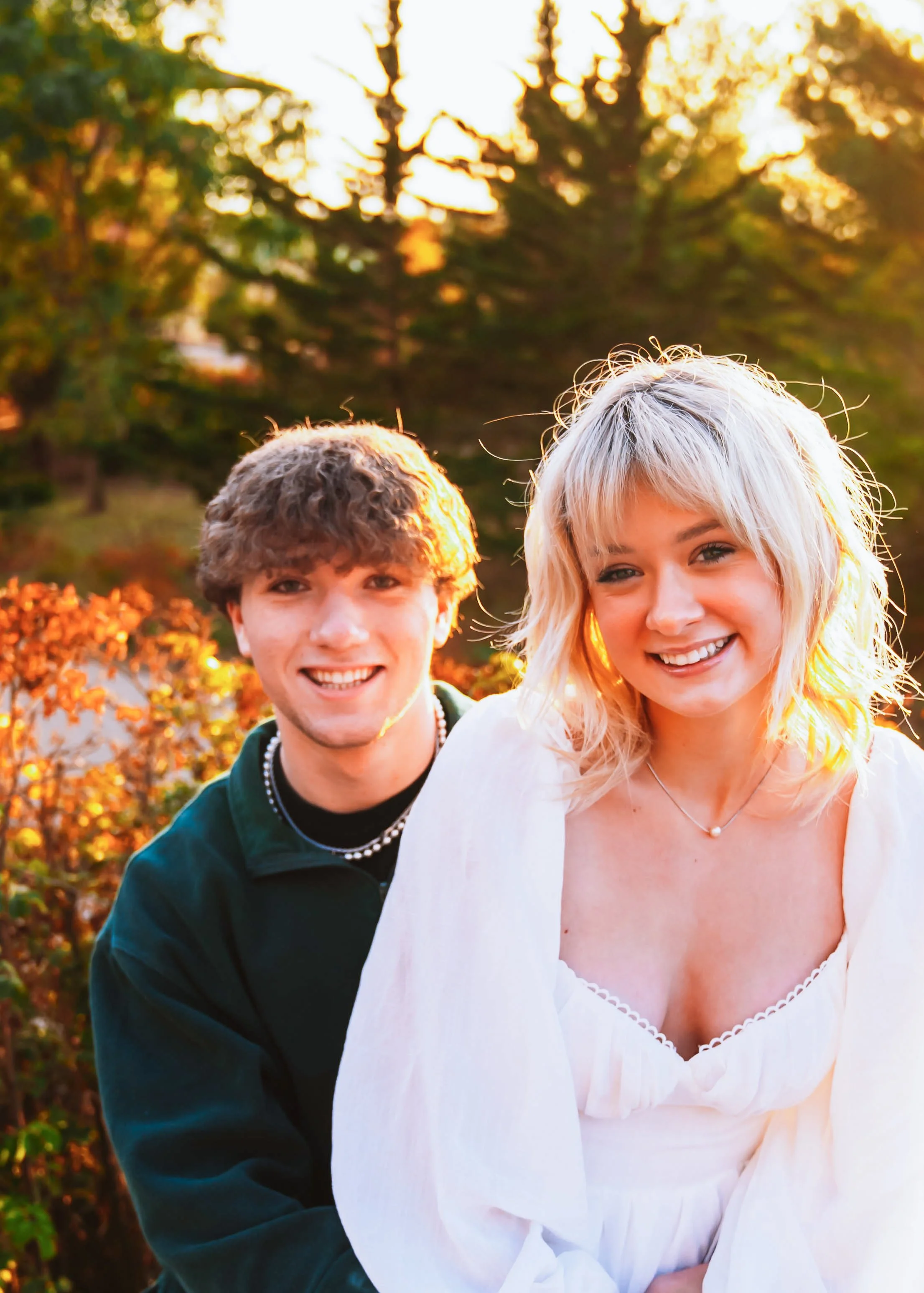 A young couple smiling outdoors during sunset, with trees and autumn foliage in the background.