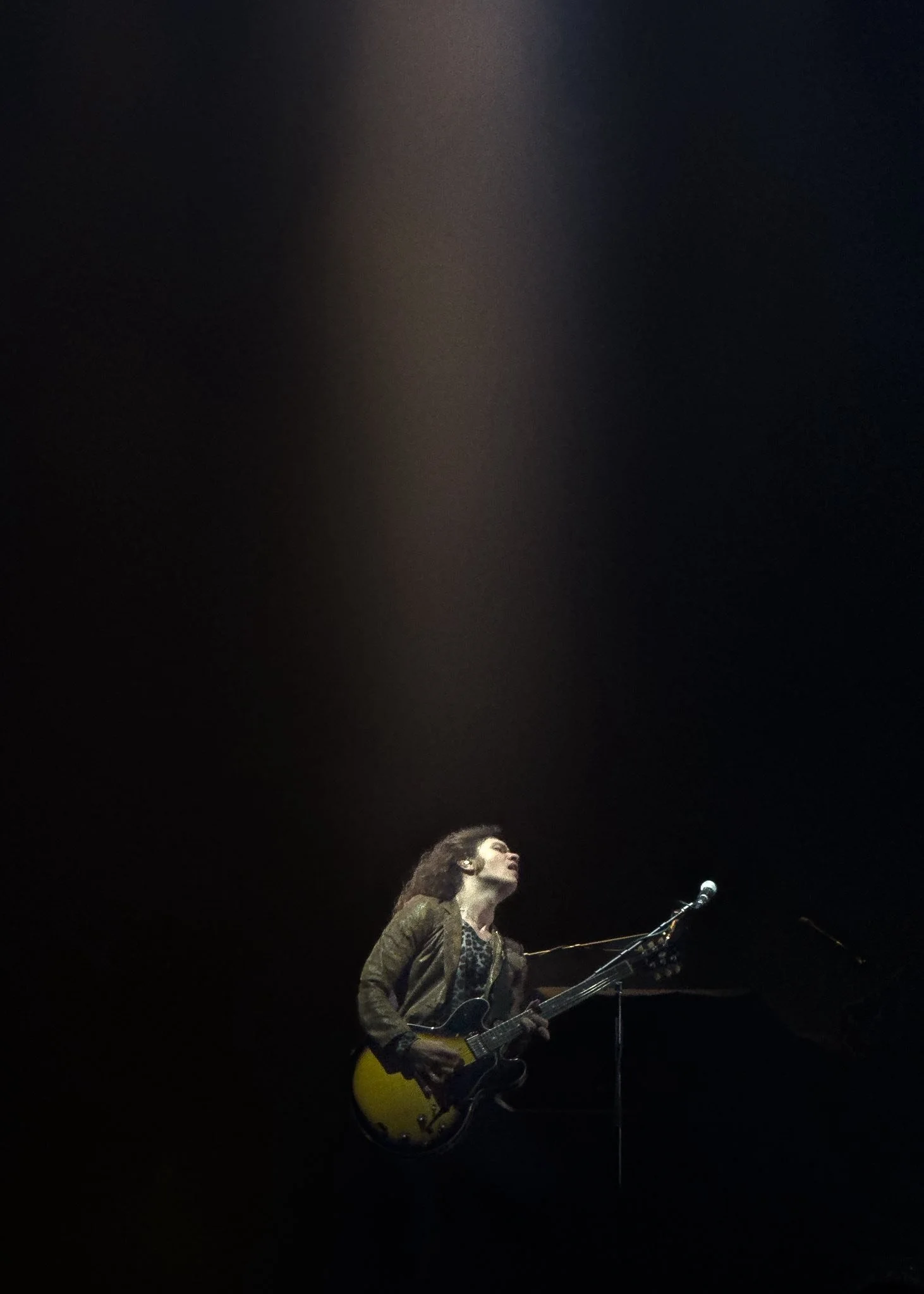 A woman with curly hair playing an electric guitar on stage, illuminated by a spotlight, with a dark background.