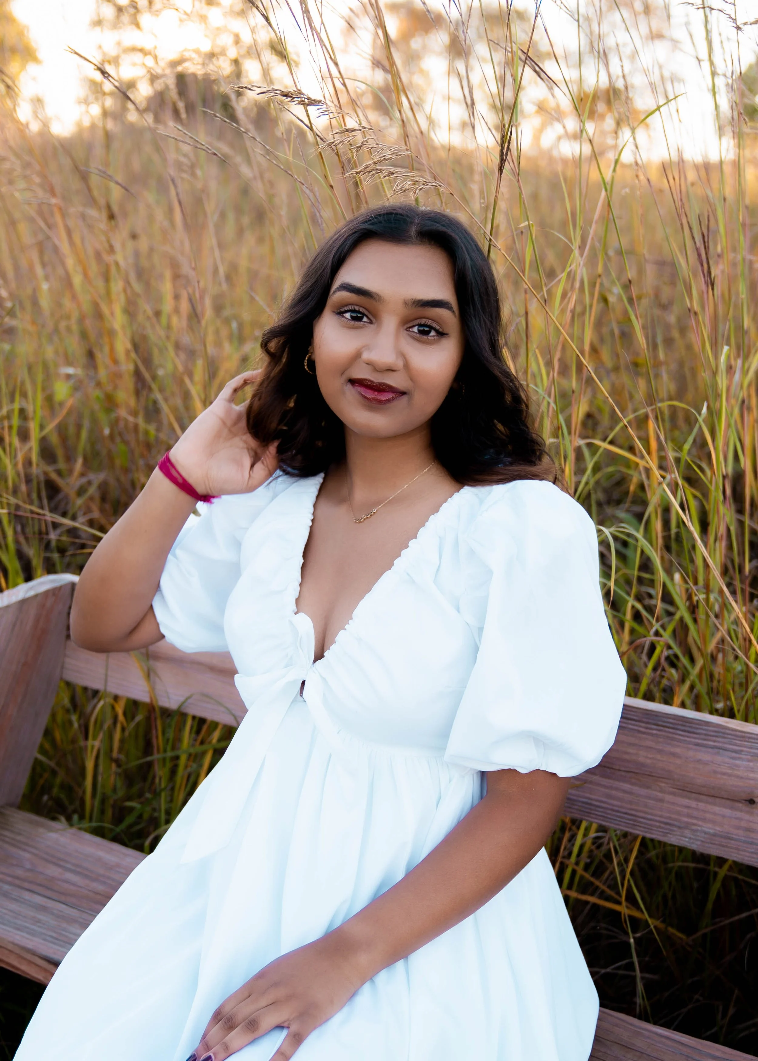 Young woman with dark hair in a white dress sitting on a wooden bench in a field of tall grass, with a sunset in the background.