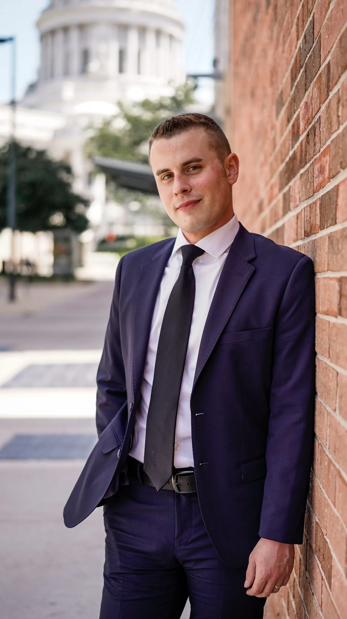 A young man in a navy suit and black tie leaning against a brick wall on city street, with a blurred building in the background.