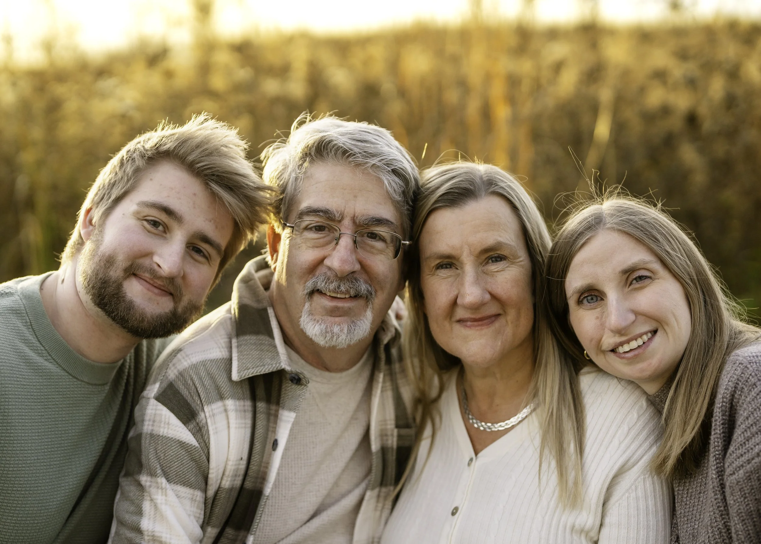 A multi-generational family of five smiling outdoors during sunset, with trees in the background.