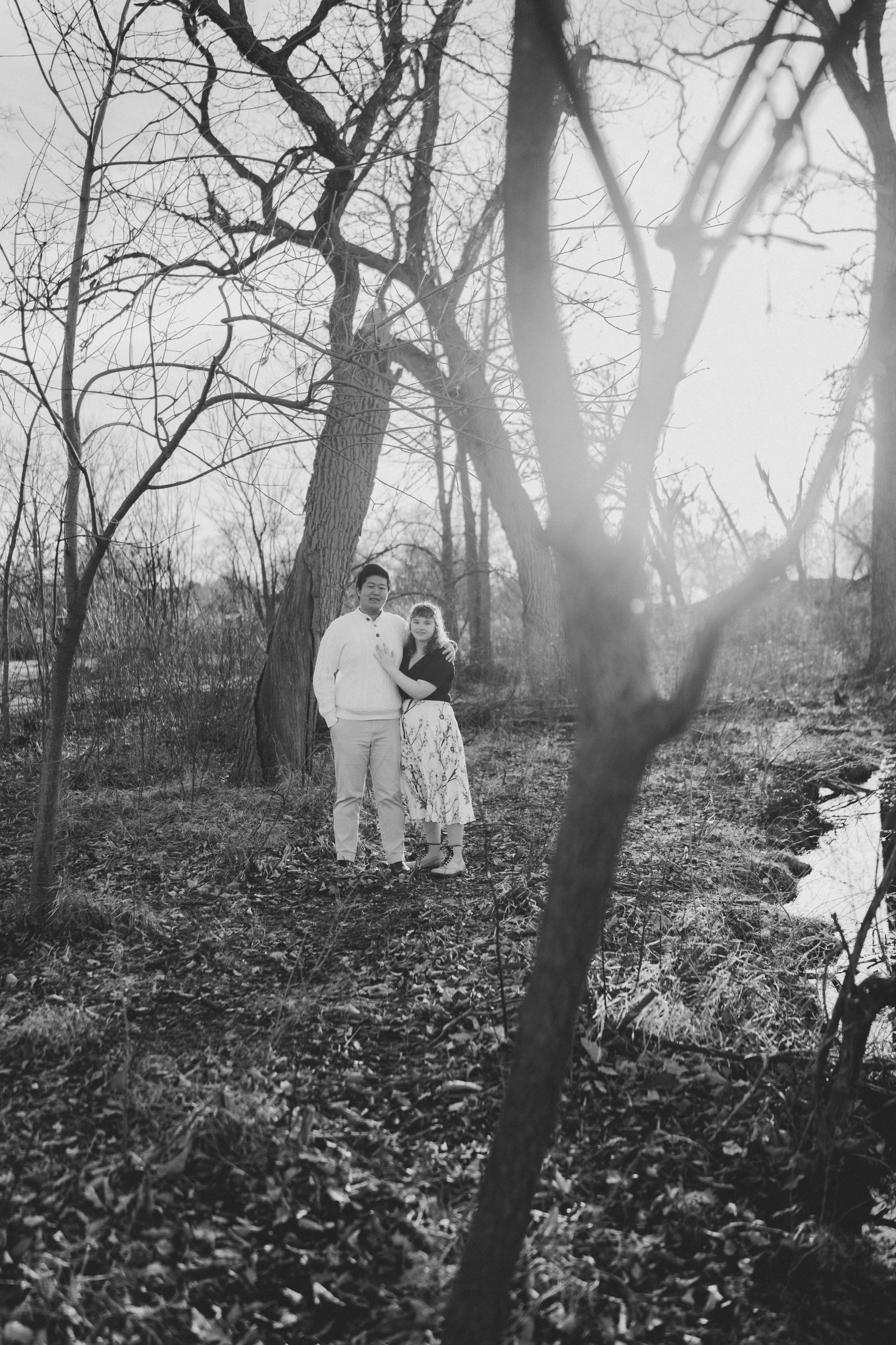 A black and white photo of a couple standing together outdoors among leafless trees near a small stream.