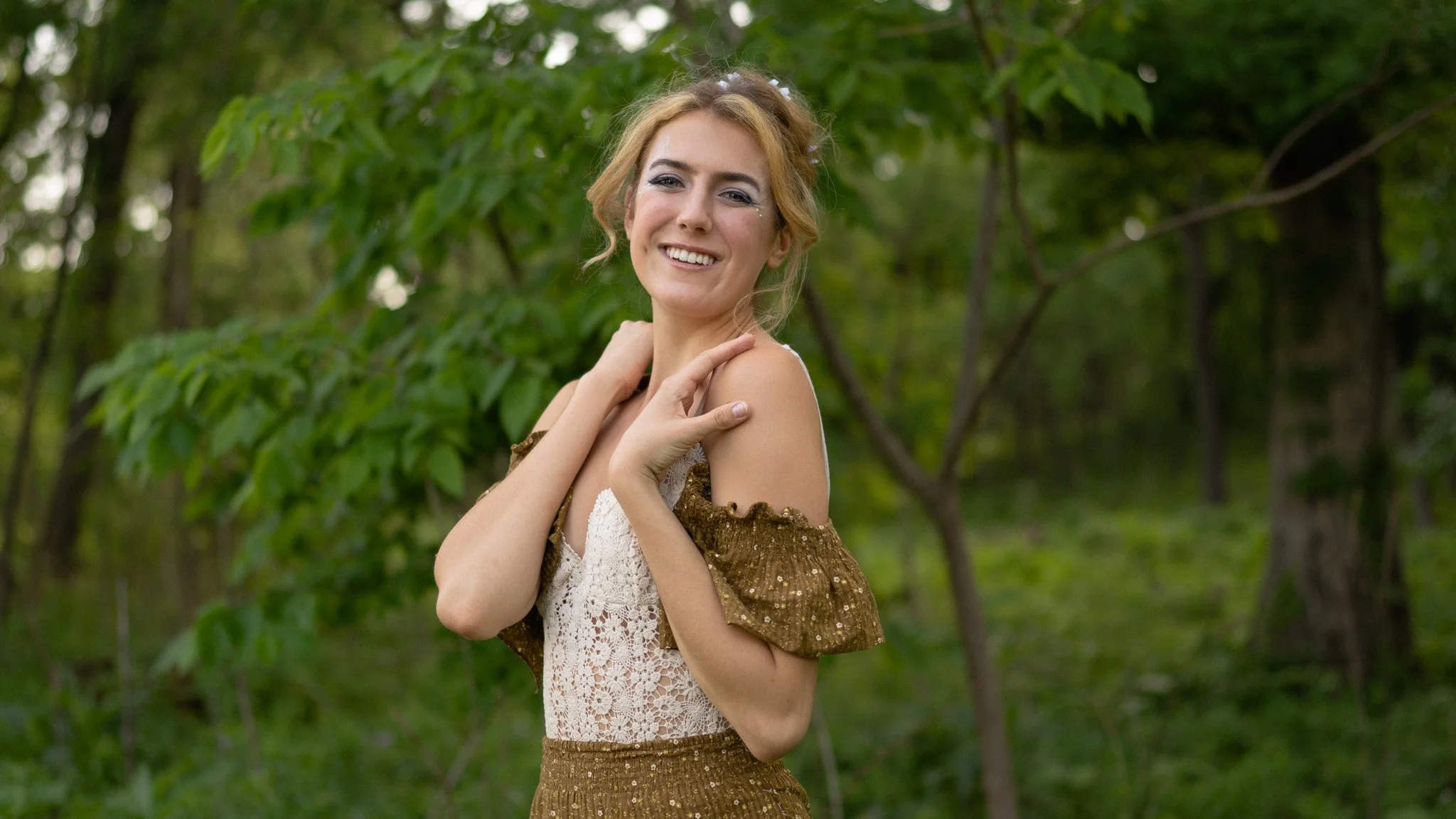 A woman with red hair smiling and touching her shoulders outdoors in a green forest.