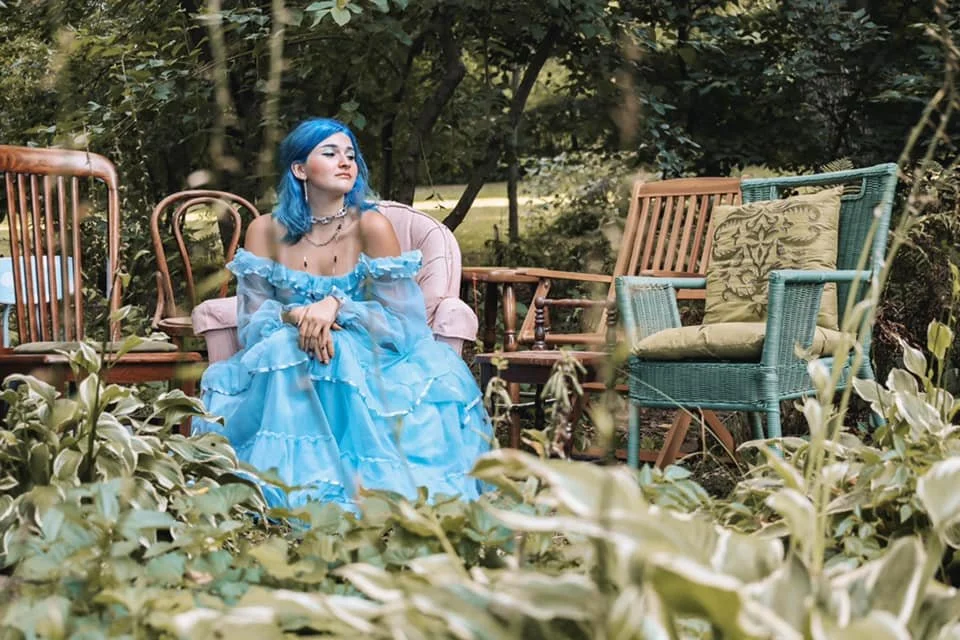 A young woman with blue hair in a light blue off-shoulder dress sitting on a pink cushioned outdoor sofa in a garden, surrounded by various chairs and lush greenery.