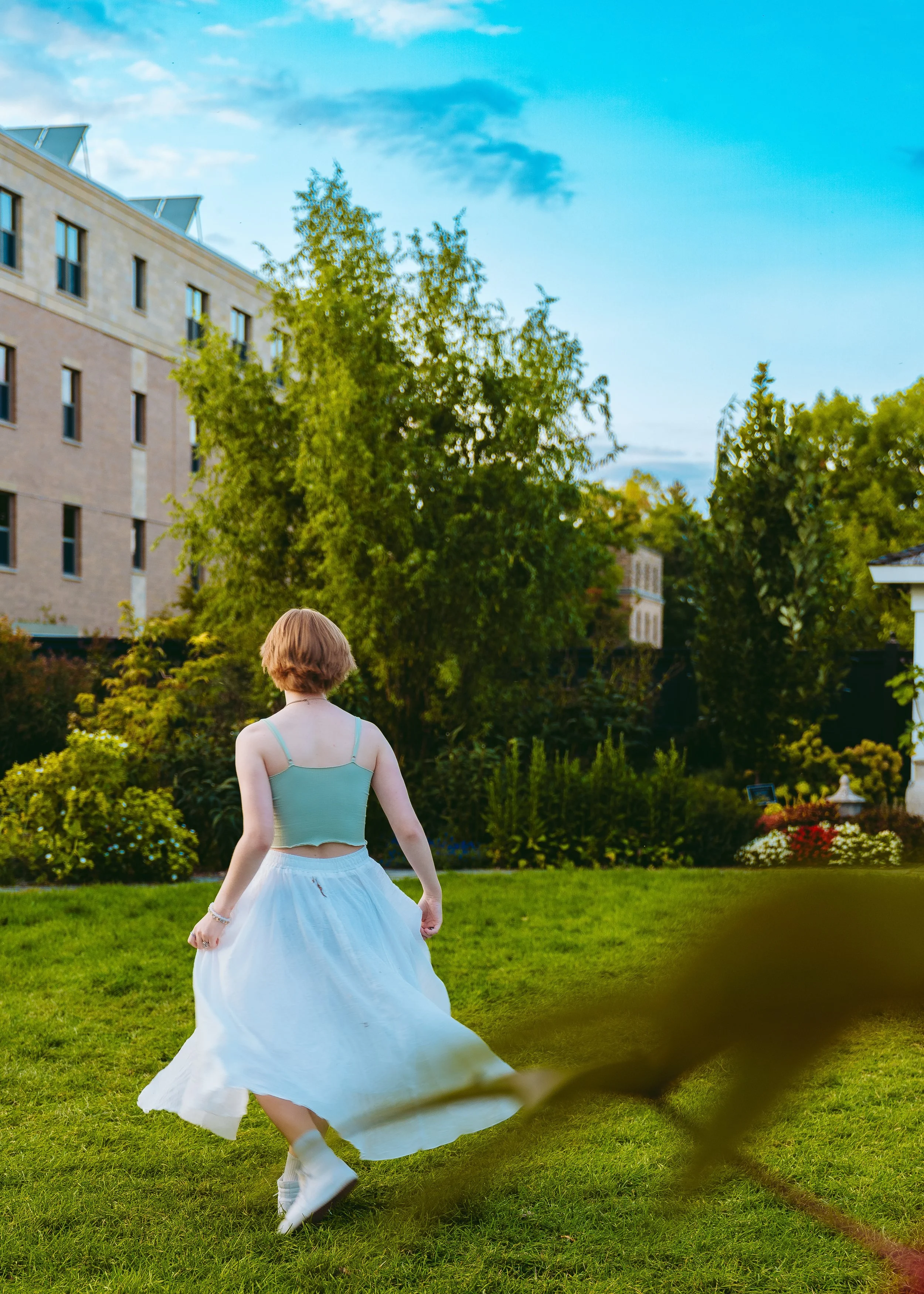 A girl with short brown hair wearing a teal spaghetti strap top and a long white skirt twirling on a green lawn in a garden with trees and flowers. There is a building in the background and a cloudy blue sky.