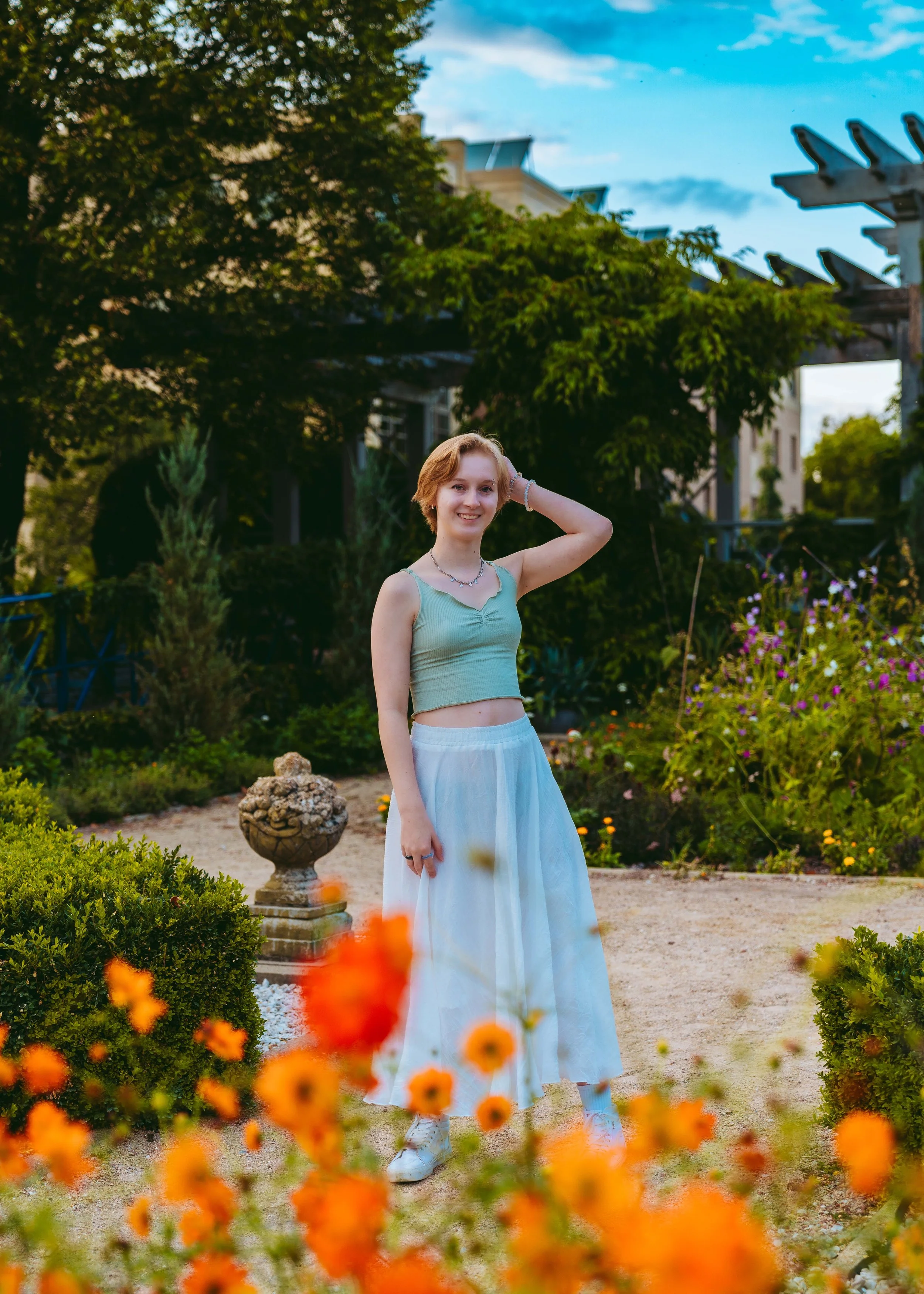 A young woman with short red hair wearing a light blue crop top and a long white skirt standing in a garden with orange flowers in the foreground, green bushes and trees in the background, and a partly cloudy sky.