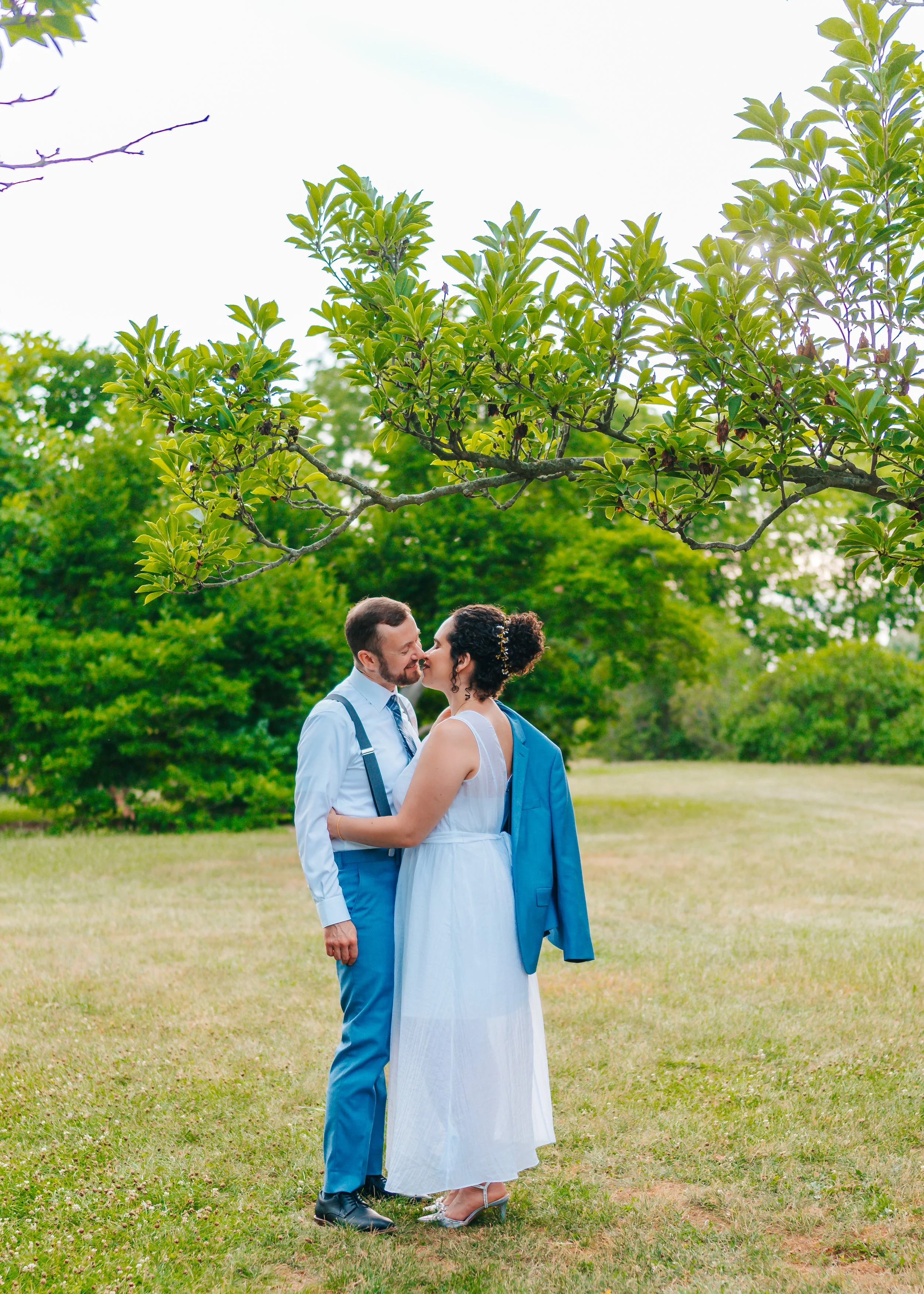 A couple dressed in formal attire sharing a kiss outdoors beneath a tree, with greenery and an open sky in the background.