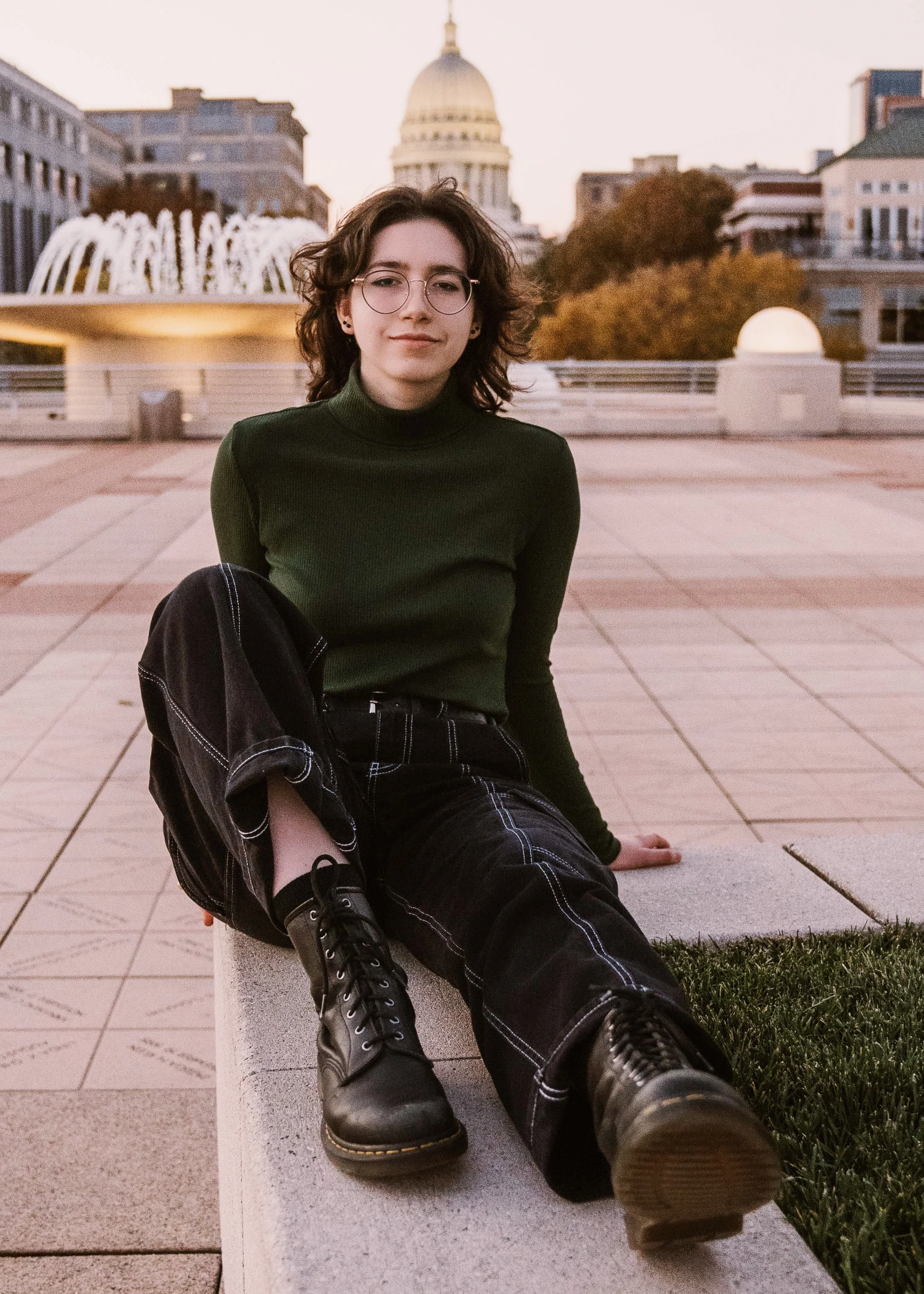 A young woman with glasses, curly hair, wearing a green turtleneck sweater and black pants with white stitching, sits on a curb in front of a fountain, with a cityscape and a domed capitol building in the background at sunset.