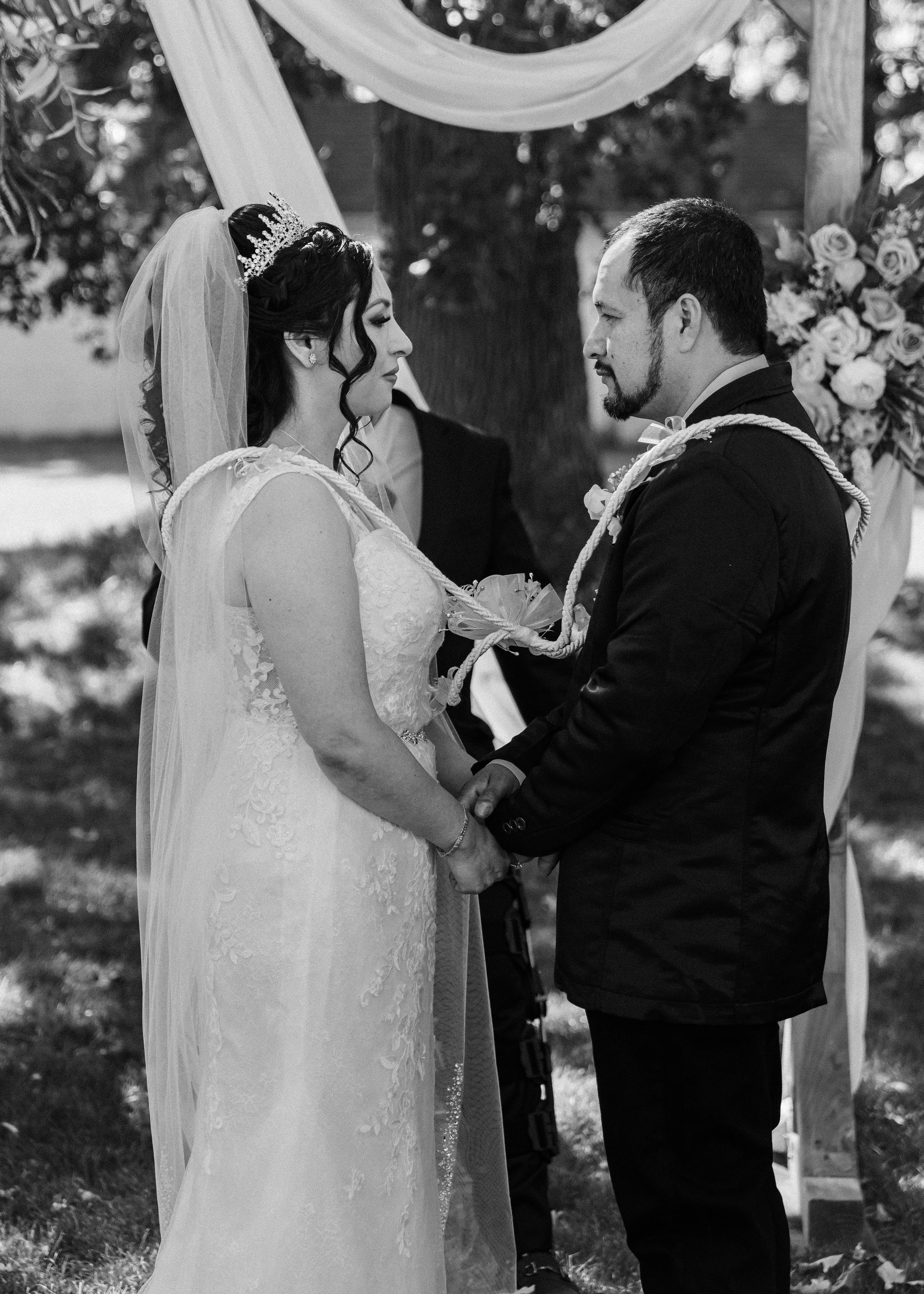 Black and white photo of a bride and groom holding hands during a wedding ceremony, standing facing each other outdoors under a wooden arch decorated with flowers and fabric, with a person officiating in the background.