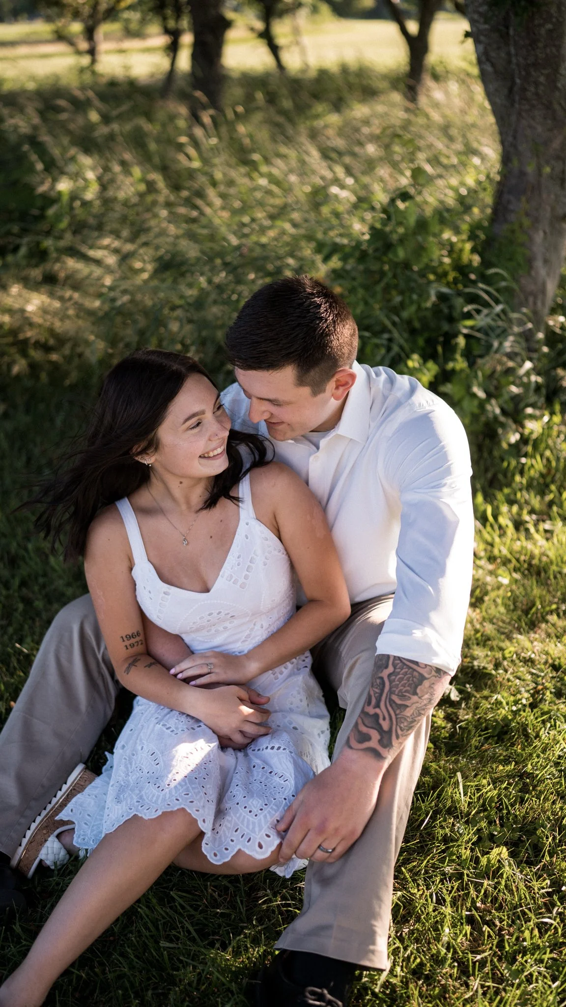 A smiling woman and a man with tattoos sitting together outdoors under a tree, enjoying a moment of affection.