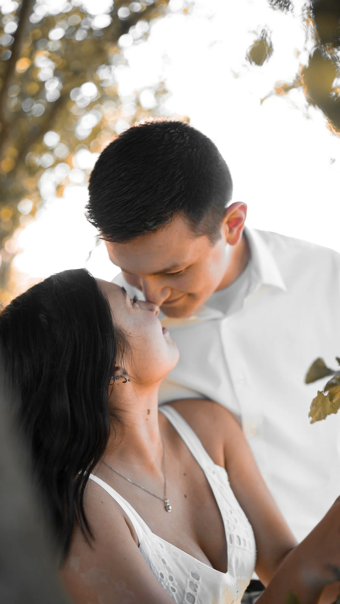 A couple sharing an intimate moment outdoors, about to kiss, surrounded by trees and warm sunlight.