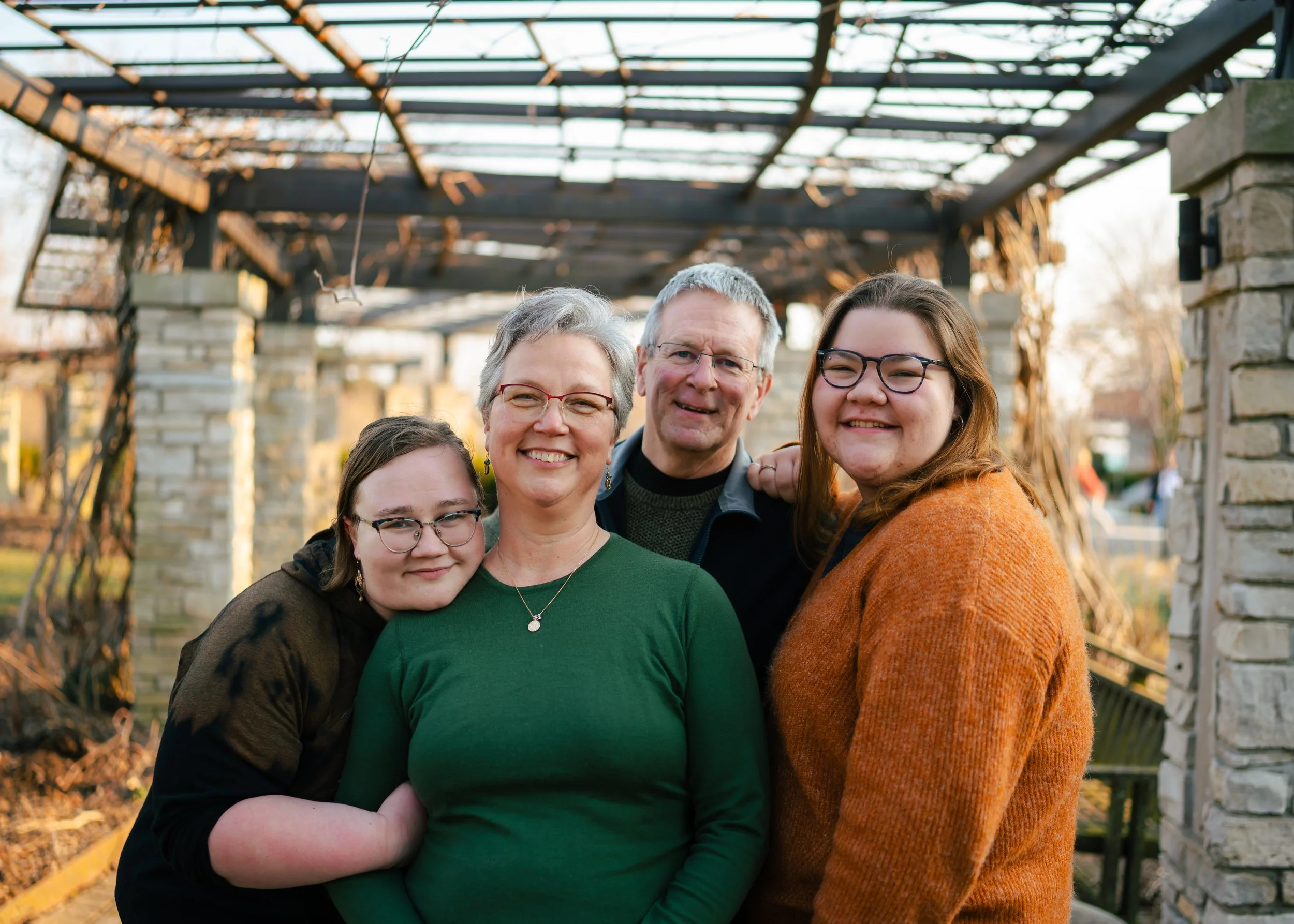 A family of five adults and teenagers smiling outdoors under an open wooden pergola with stone columns, in a garden or park during late afternoon.