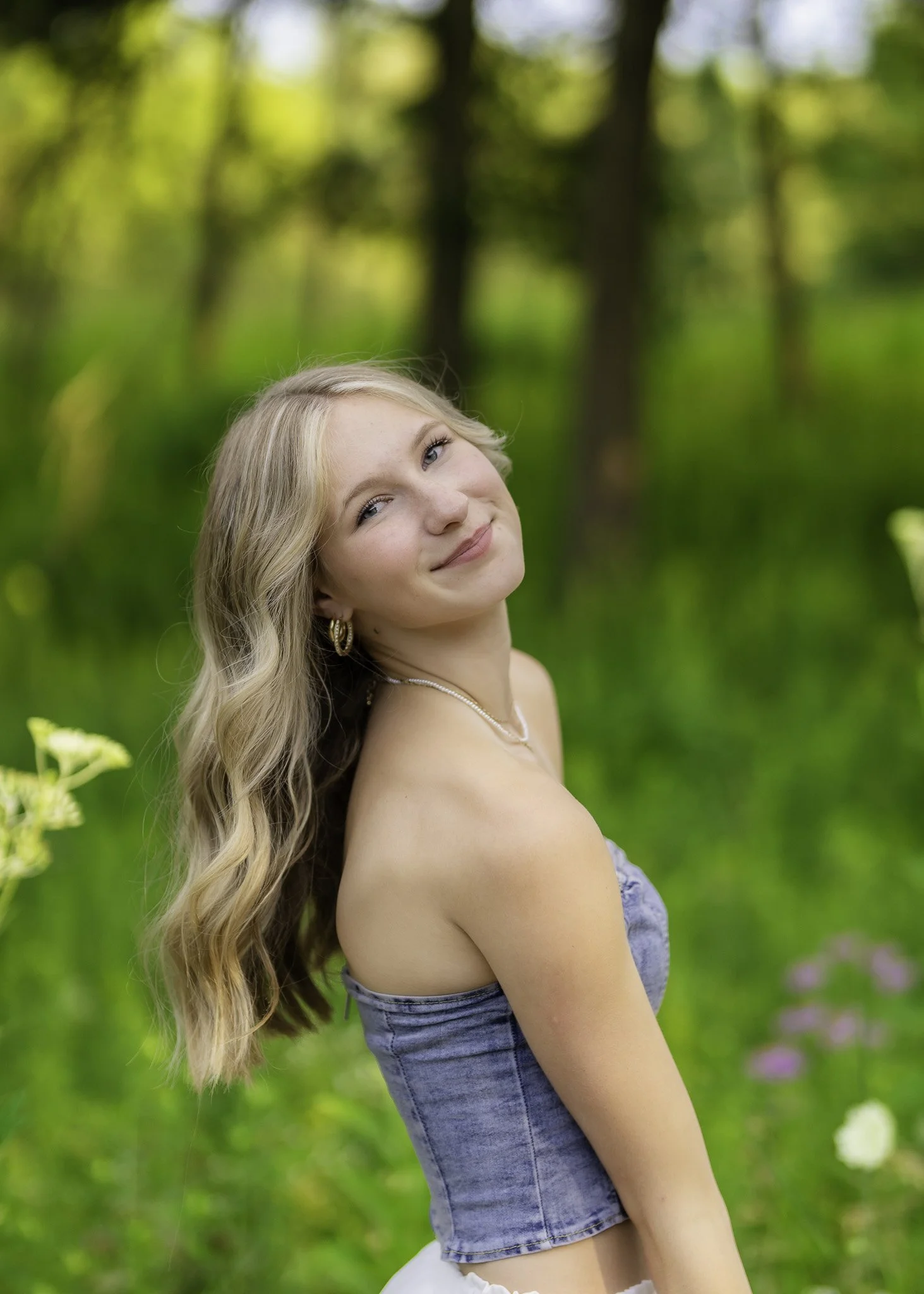 A young woman with long blonde hair, wearing a strapless denim top and jewelry, smiling in a green outdoor setting with trees and flowers in the background.