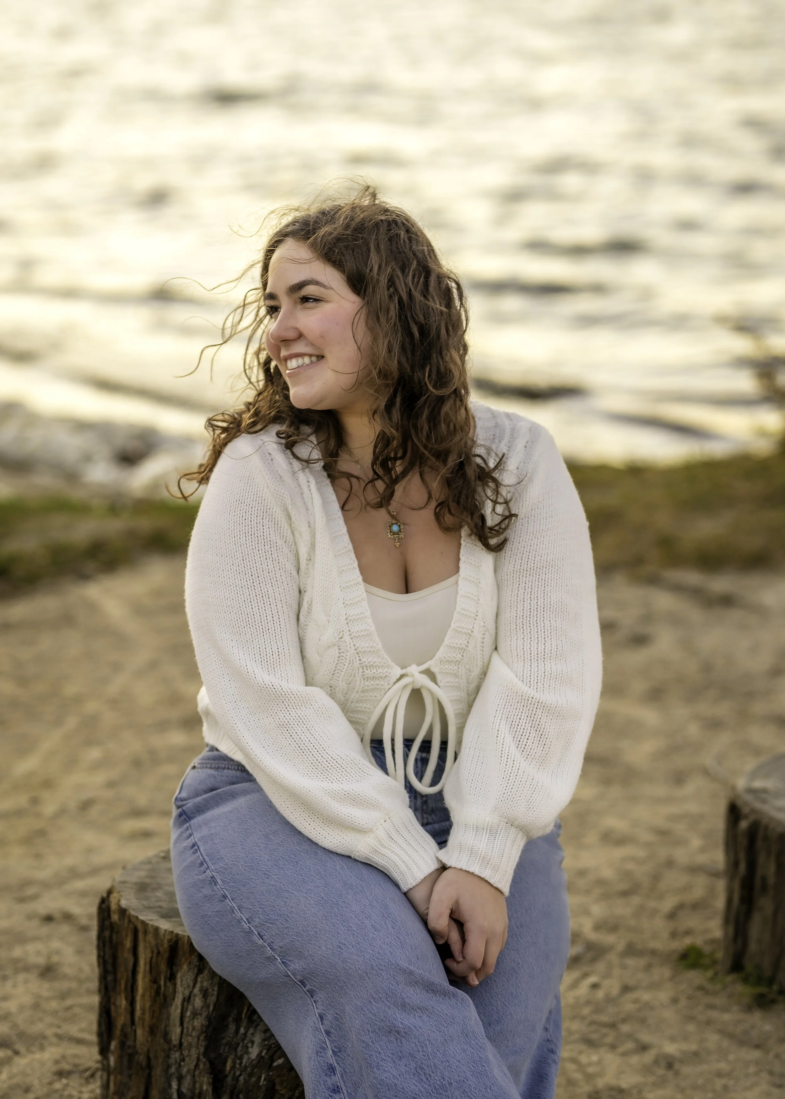A young woman with curly brown hair smiling while sitting on a tree stump near a body of water during sunset.