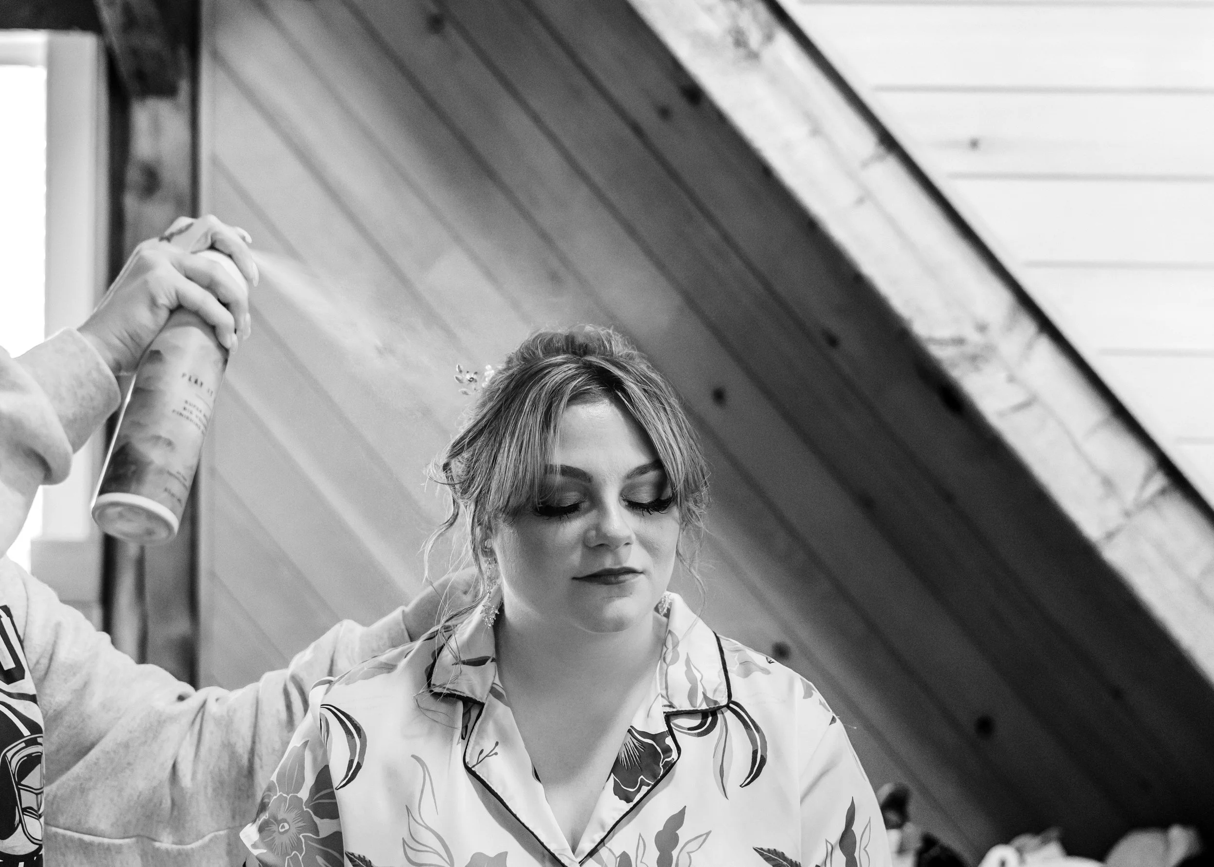 A woman with styled hair and makeup sitting with eyes closed, while a person sprays her hair with a can of hair spray in a room with wooden panel ceiling.