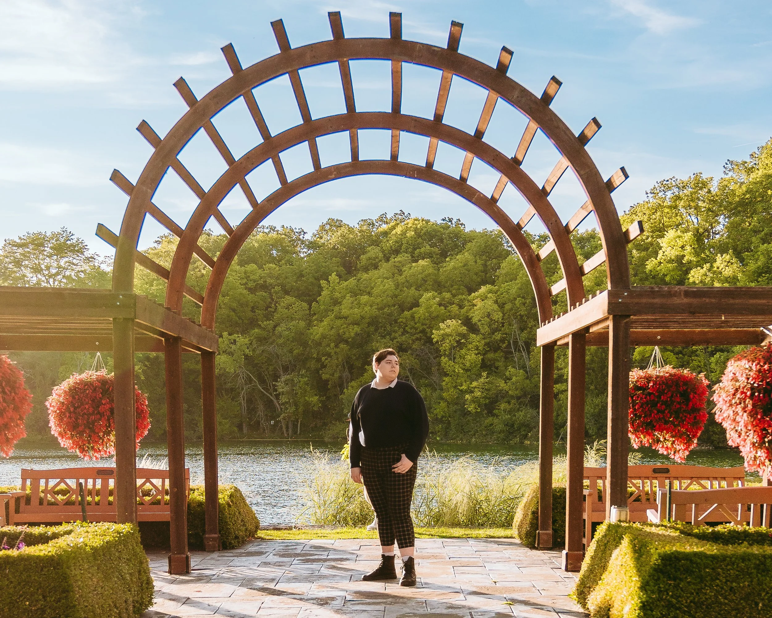 A person standing in front of a lakeside wooden arbor with hanging flower baskets, lush green trees in the background, and a clear blue sky.
