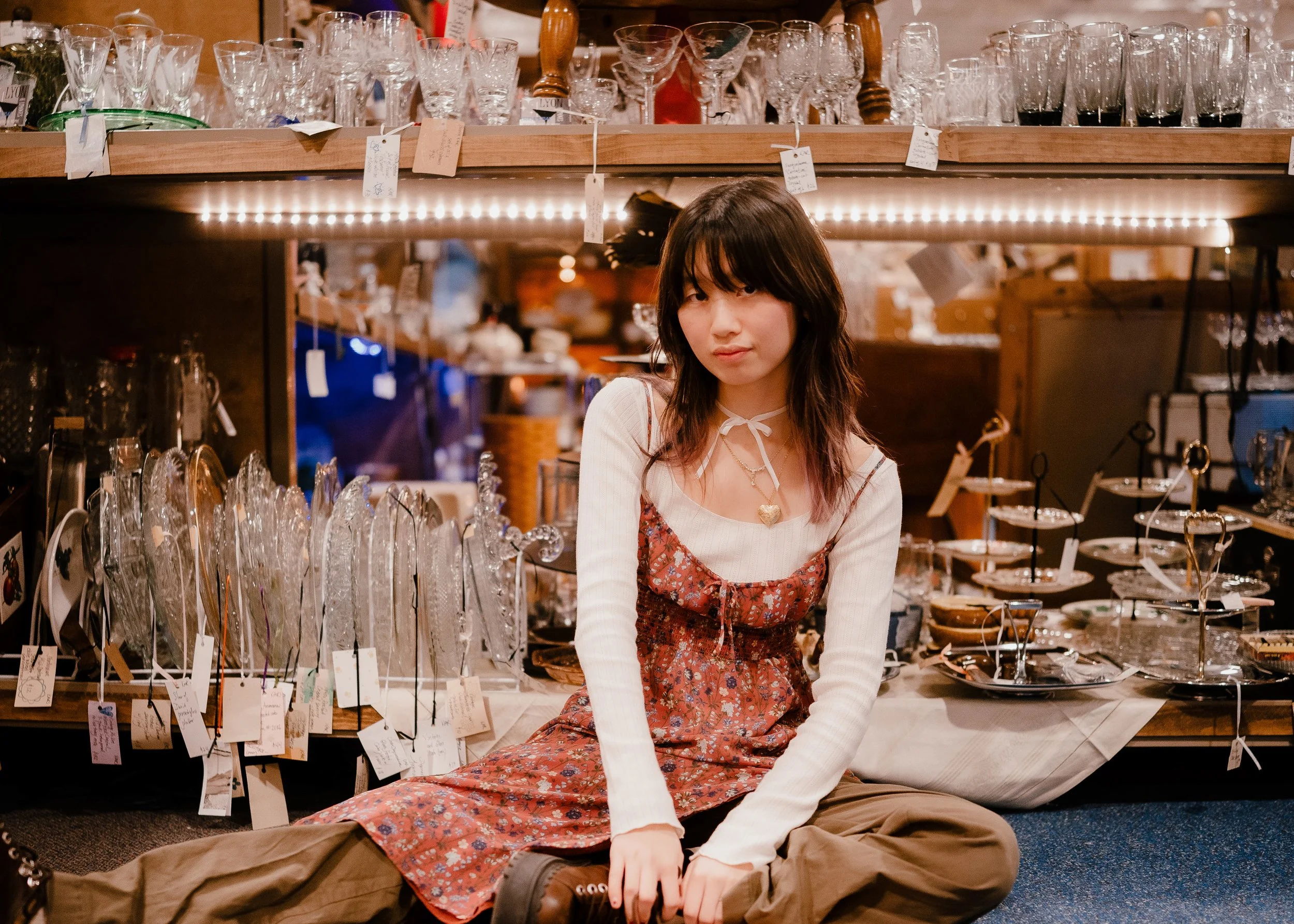 A young woman with dark hair sitting on the floor in front of a display of glassware at an antique shop or market. She is wearing a layered dress, a cream long sleeve shirt, and has a heart necklace. The background shows shelves with various glass an