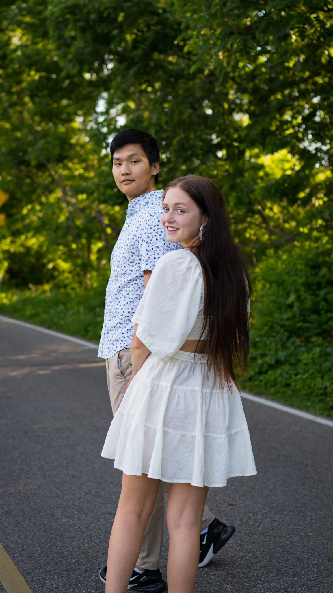 A young man and a young woman standing on a road surrounded by green trees. The woman is smiling and wearing a white dress with long dark hair. The man is standing slightly behind her, wearing a patterned short sleeve shirt and beige pants, looking t