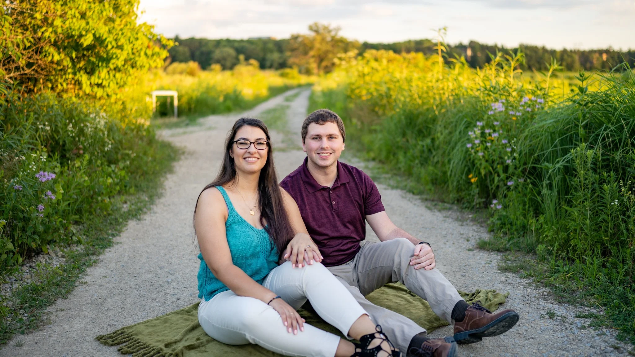 A young couple sitting on a blanket on a country dirt road, smiling at the camera, surrounded by greenery and wildflowers in a rural setting during late afternoon.