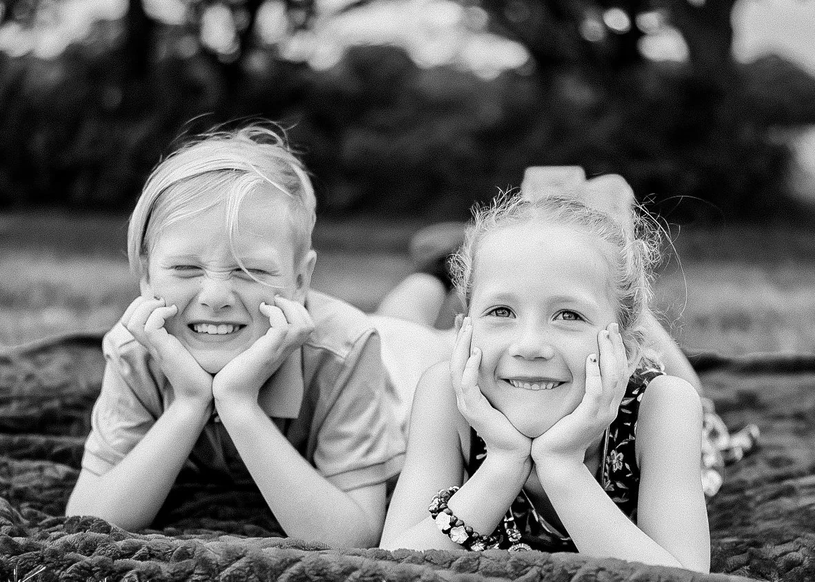 Two children, a boy and a girl, lying on their stomachs on a blanket, resting their chins on their hands, and smiling at the camera. The boy is on the left with short, light-colored hair, and the girl is on the right with long, curly hair tied back.