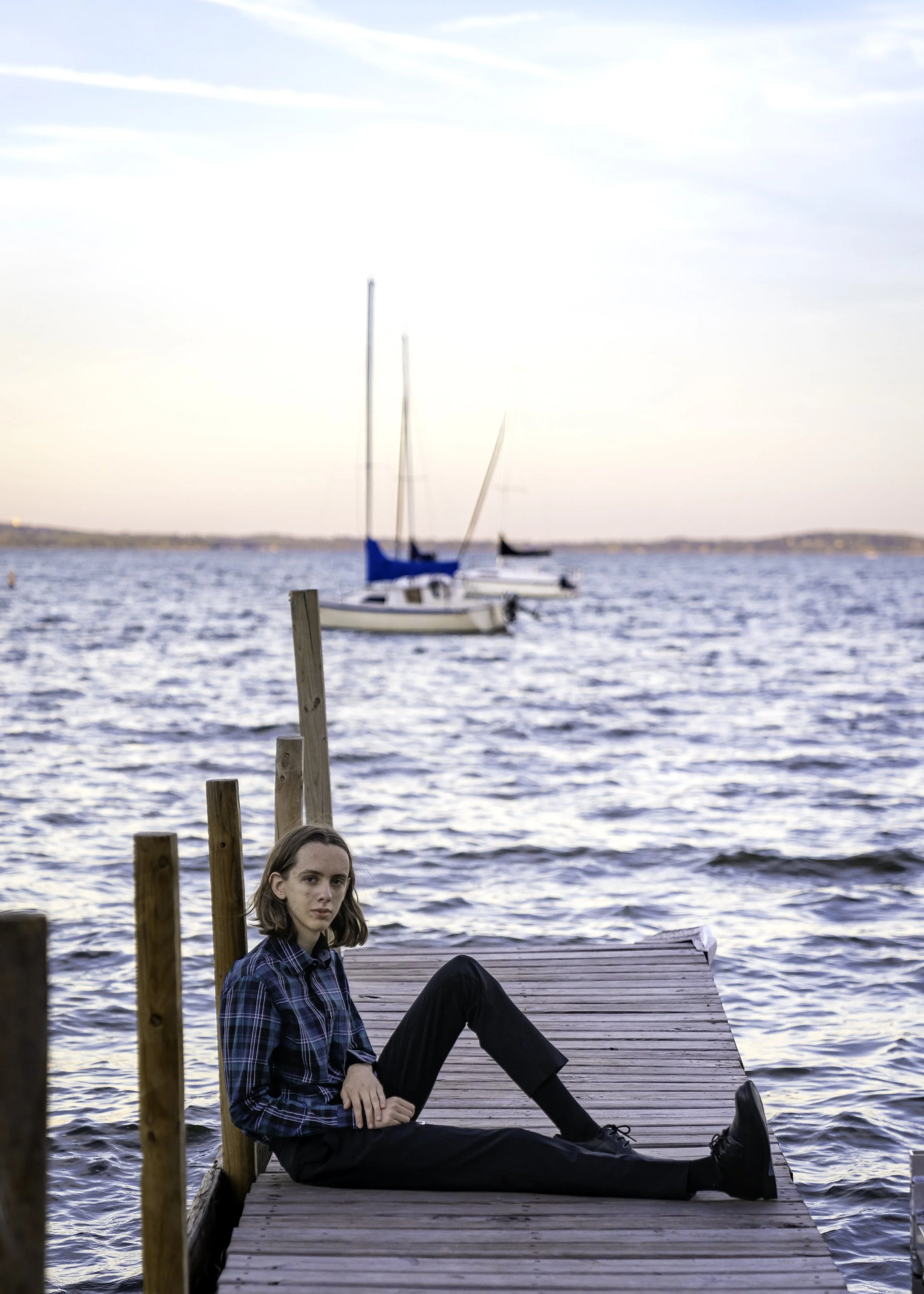 A young woman with shoulder-length brown hair, wearing a blue plaid shirt and black pants, sitting on a wooden dock near a body of water. In the background, there are sailboats floating on the water, with a pastel-colored sky during sunset or sunrise