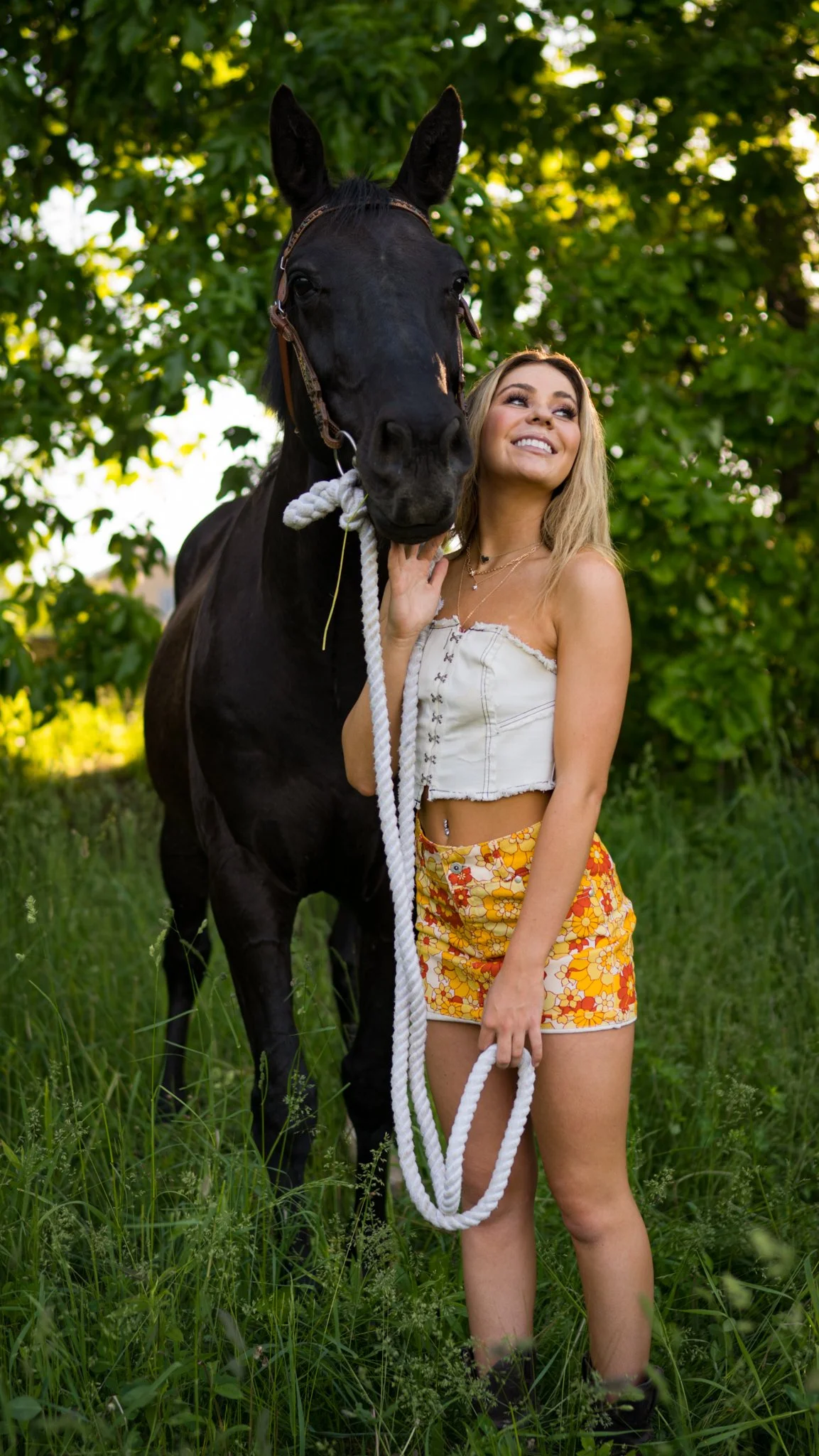 A young woman with blonde hair, wearing a white off-shoulder top and colorful shorts, stands in a grassy outdoor area, smiling and holding a white rope lead attached to a large black horse. The horse has a bridle and is standing close to her, with le