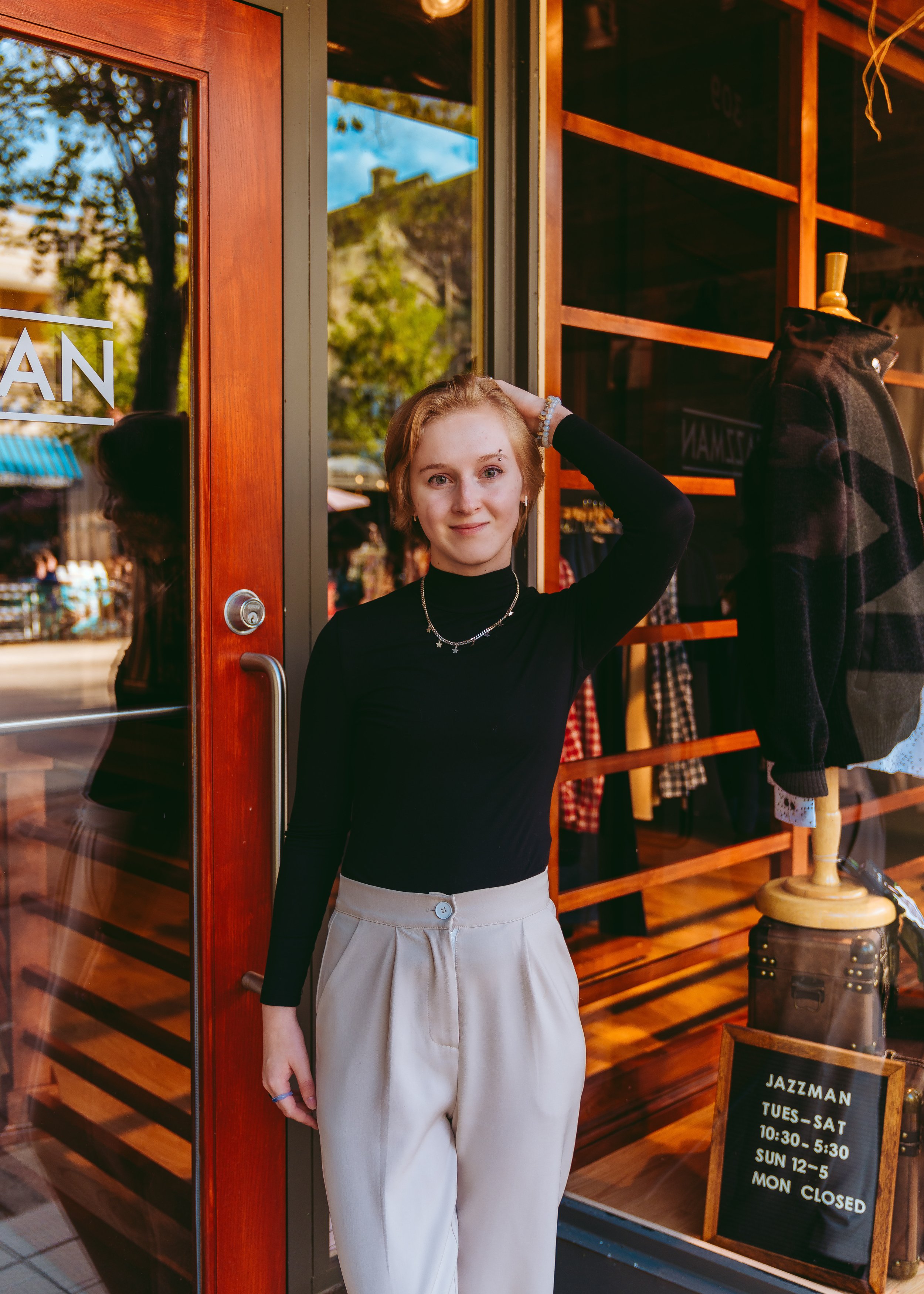 A young woman with short blonde hair dressed in a black turtleneck and beige high-waisted pants standing in front of a storefront with a glass door. She has jewelry including necklaces and a bracelet. The store’s window displays clothing and a sign w