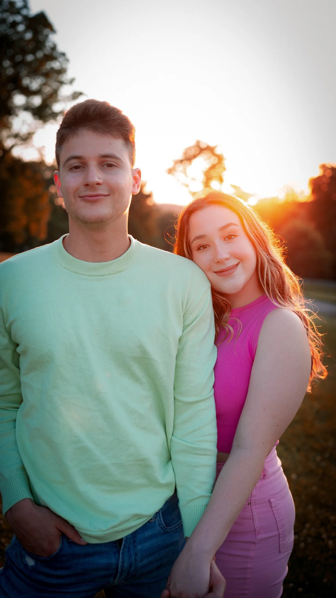 A smiling young man and woman holding hands outdoors at sunset. The man wears a light green sweater and jeans, and the woman wears a pink sleeveless top and pink pants. Trees and the setting sun are in the background.