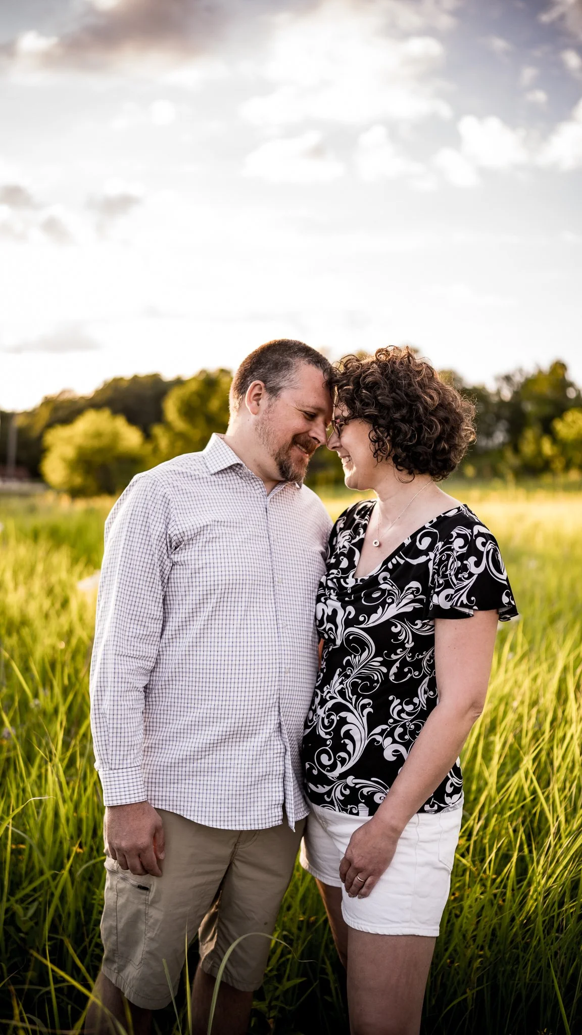A man and woman standing close together in a grassy field, touching foreheads and smiling. The man is wearing a checkered shirt and khakis, and the woman is wearing a black and white patterned top and white shorts. The background features trees and a