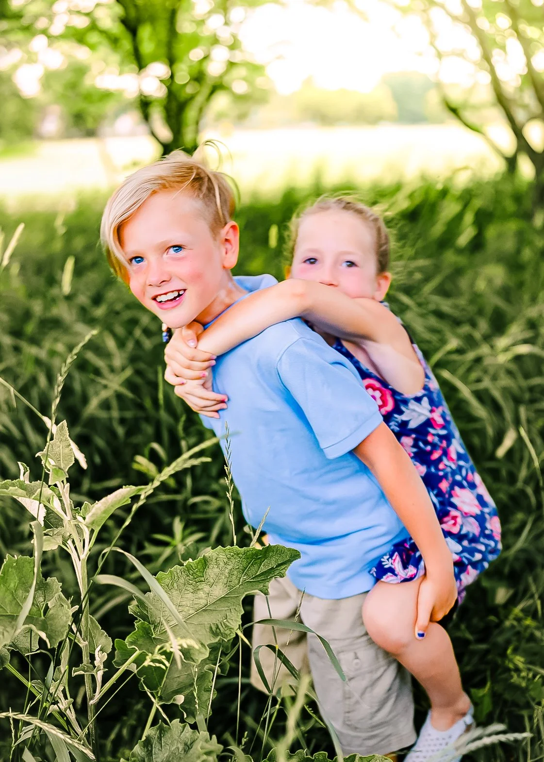 A young boy carries a girl on his back in a green field with trees in the background, both smiling and enjoying the outdoors.