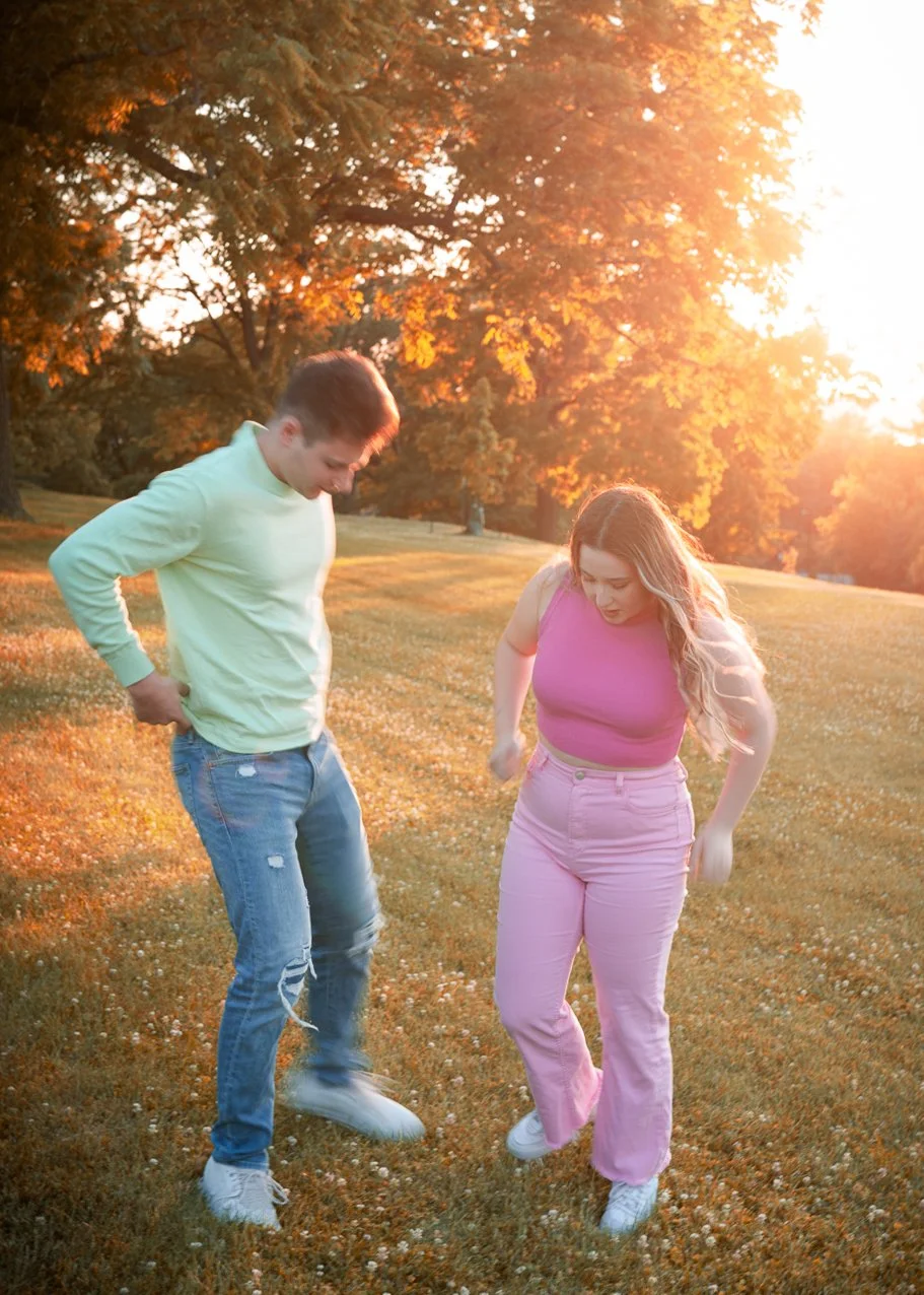 Two young people, a man and a woman, dancing or jumping in a park during sunset with orange and yellow autumn leaves on trees.