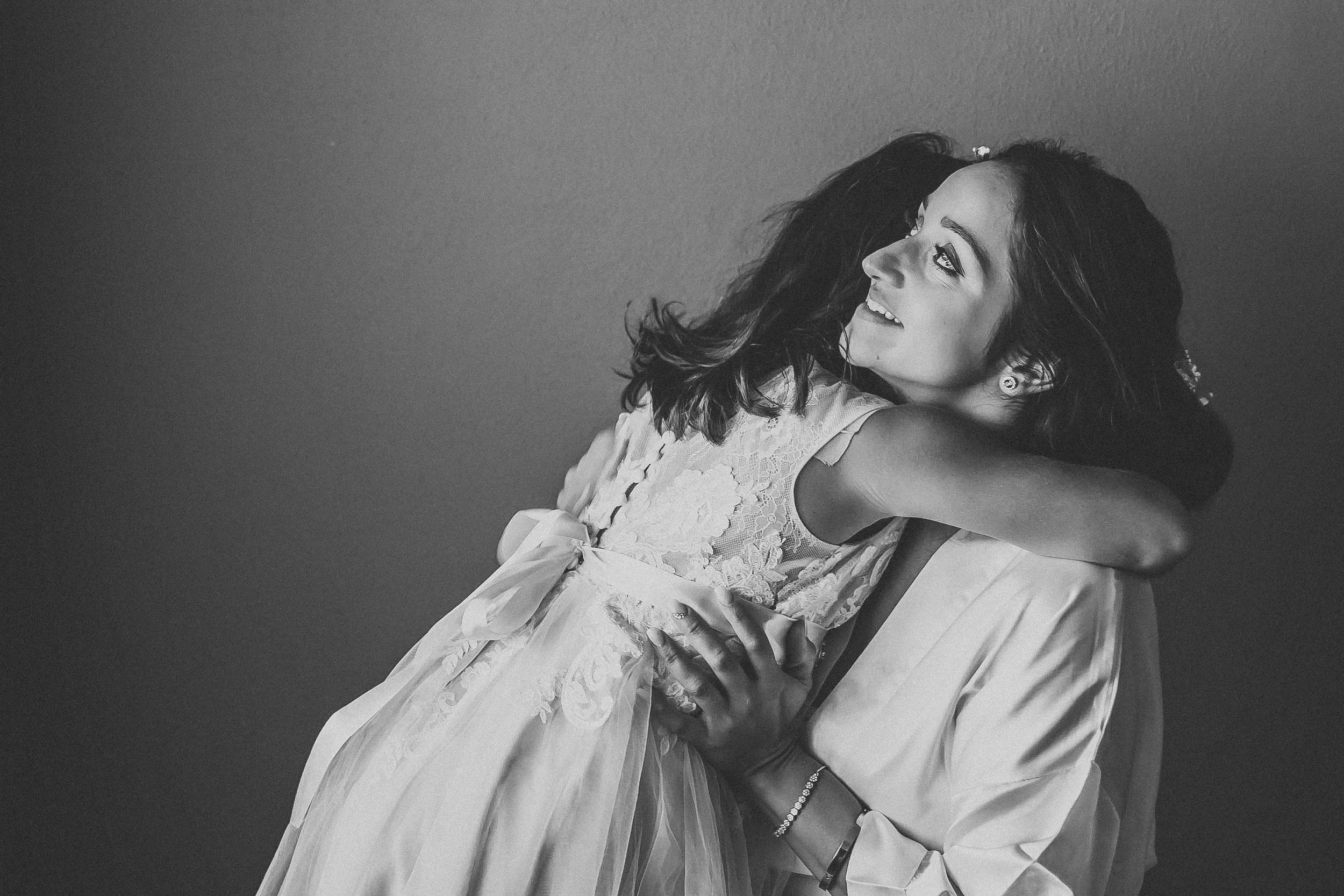 A woman in a wedding dress being hugged by a person in a suit, both smiling and looking at each other.