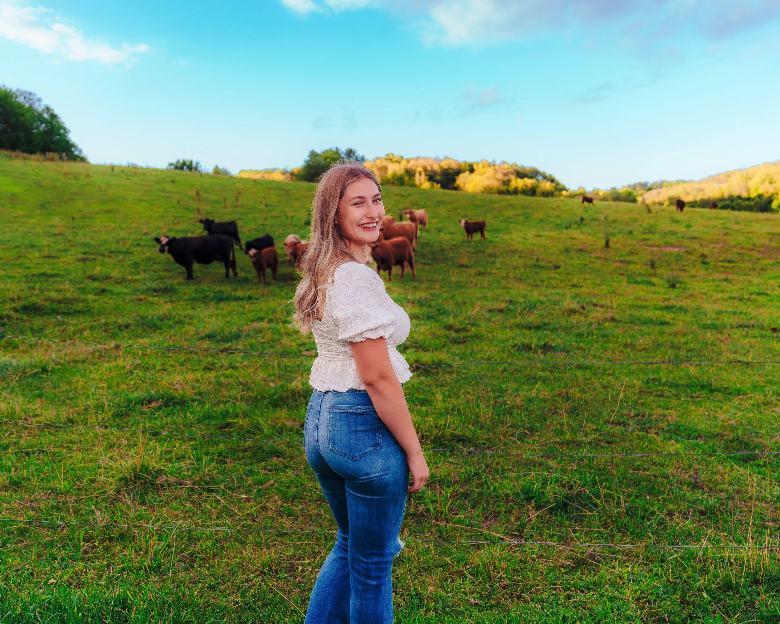 A woman standing on a grassy field near a fence, smiling, with cows grazing in the background across a hill and a partly cloudy sky.