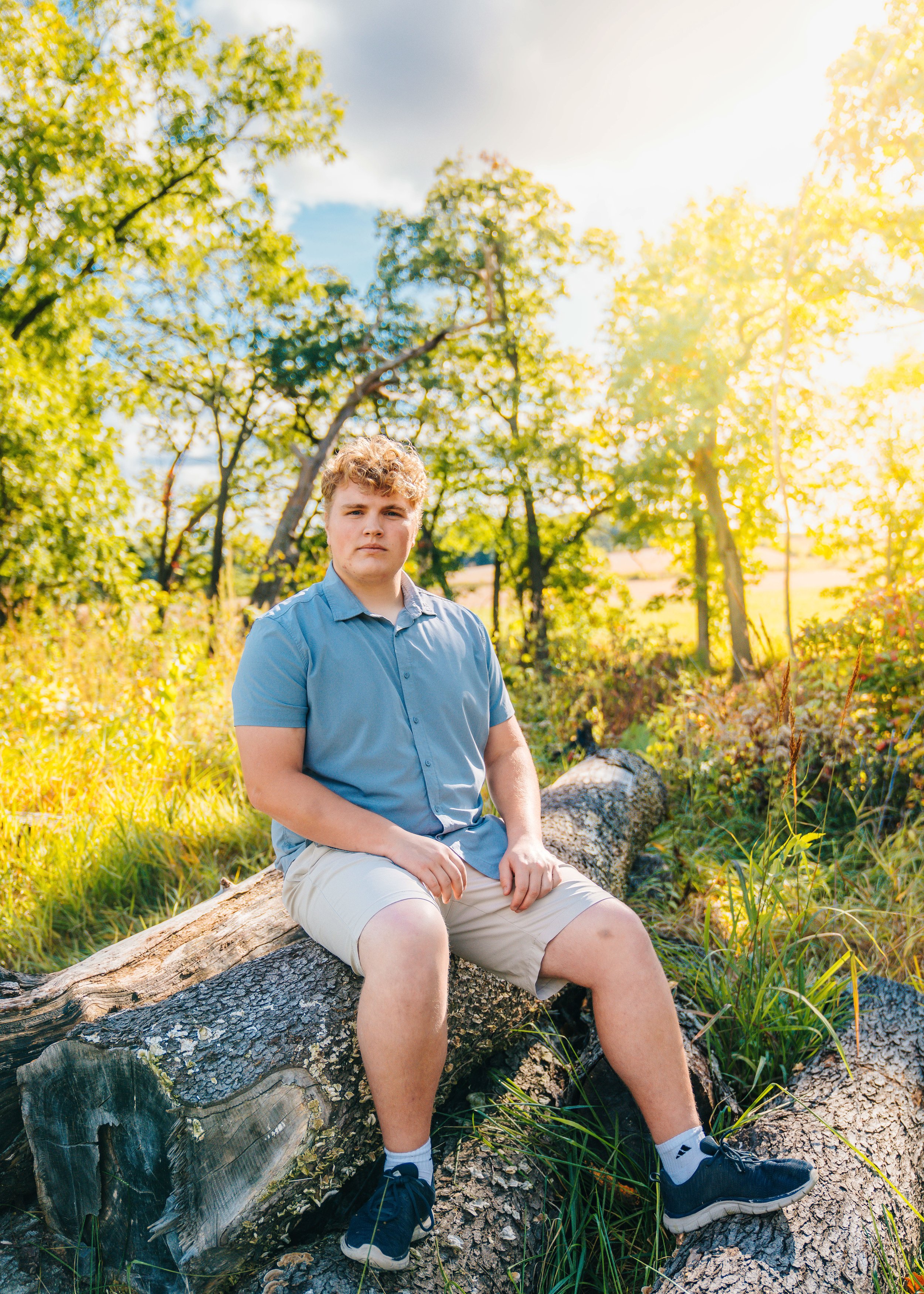 A teenage boy with curly hair sitting on a fallen tree trunk in a sunlit, green outdoor setting with trees and grass, wearing a light blue short-sleeve shirt, beige shorts, and black sneakers.