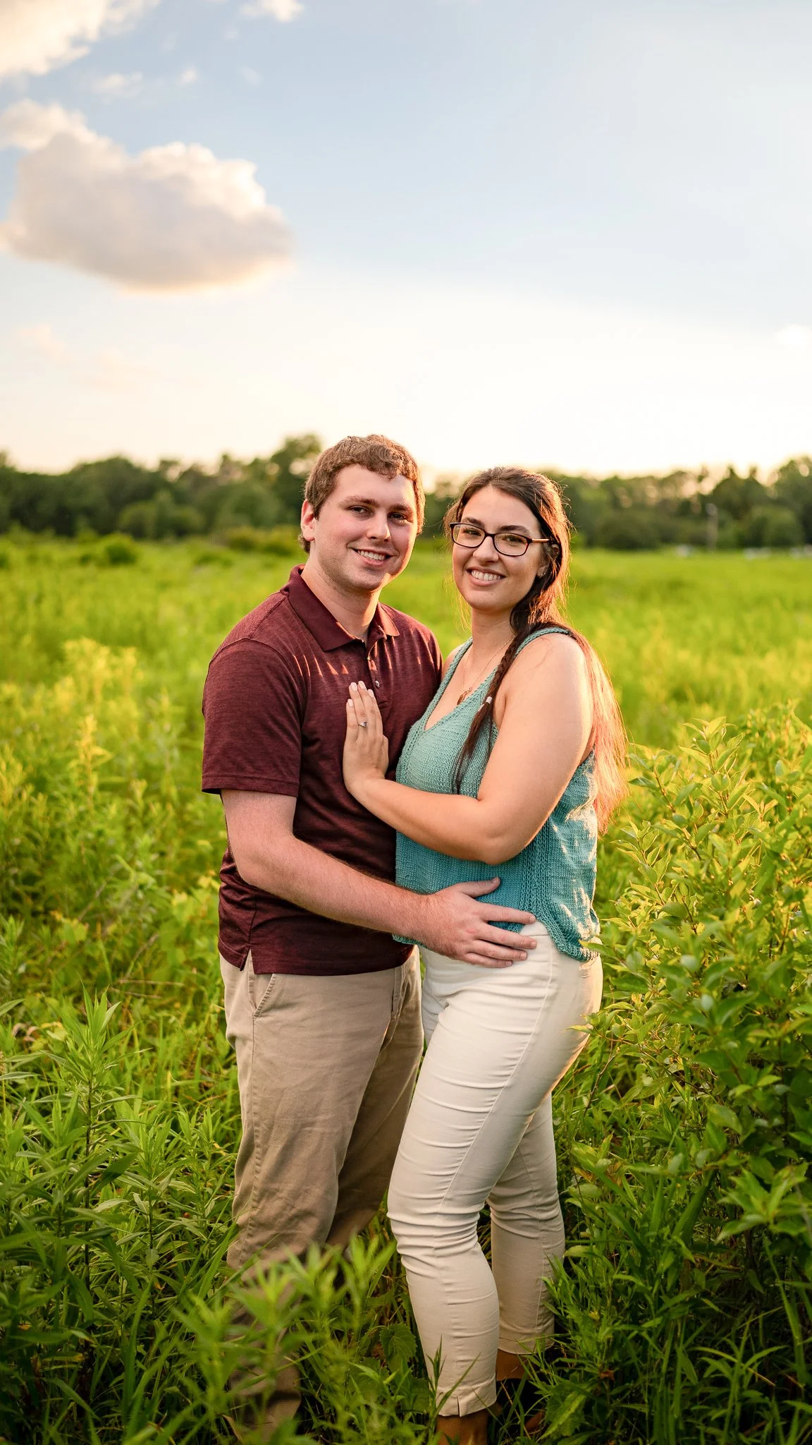 A happy couple standing close together in a green field at sunset, smiling at the camera.