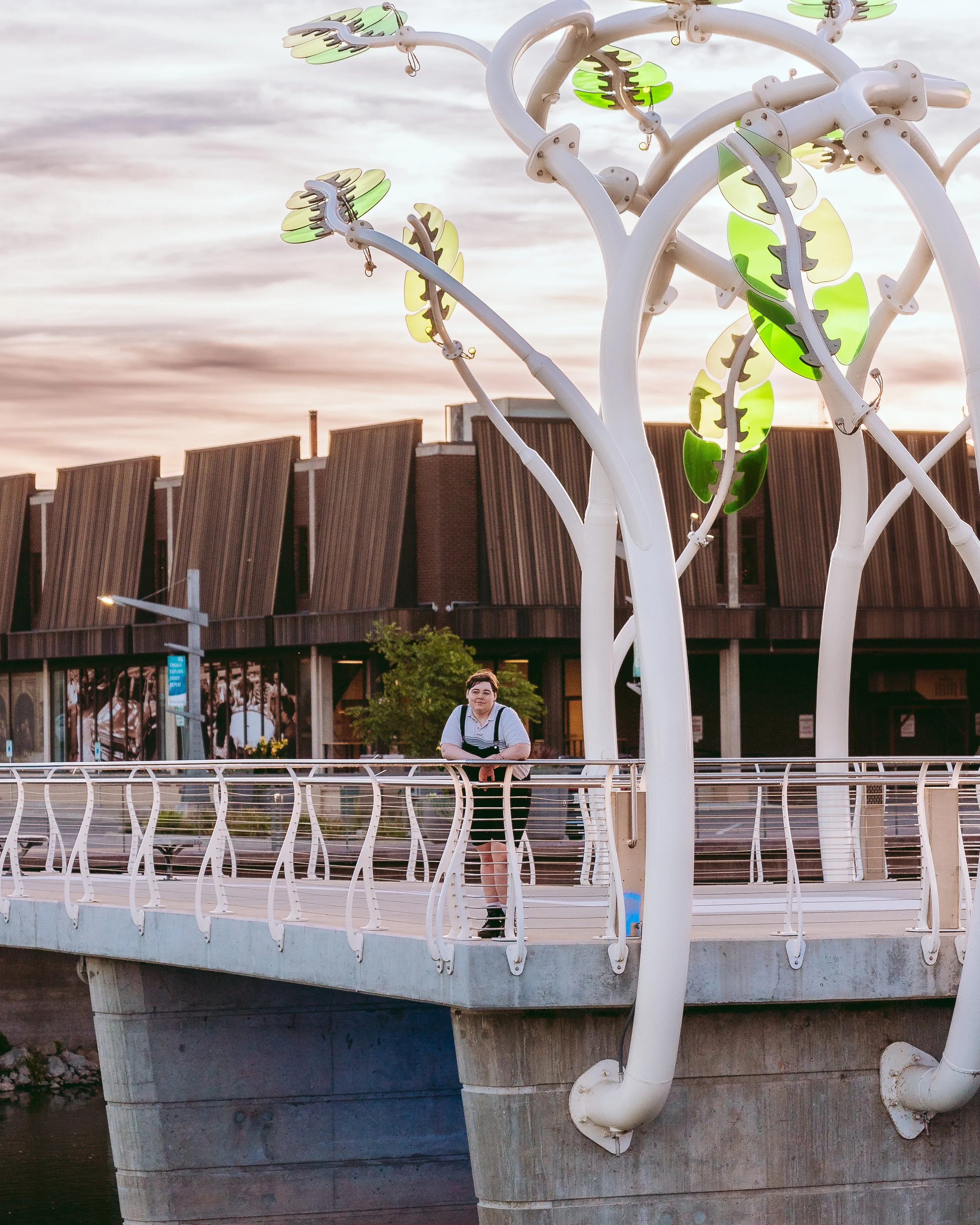 A young person stands on a bridge with modern railing, leaning on it, near decorative street lamps shaped like trees with leaves. Behind them, there is a building with unique angled brown panels and a sunset sky.