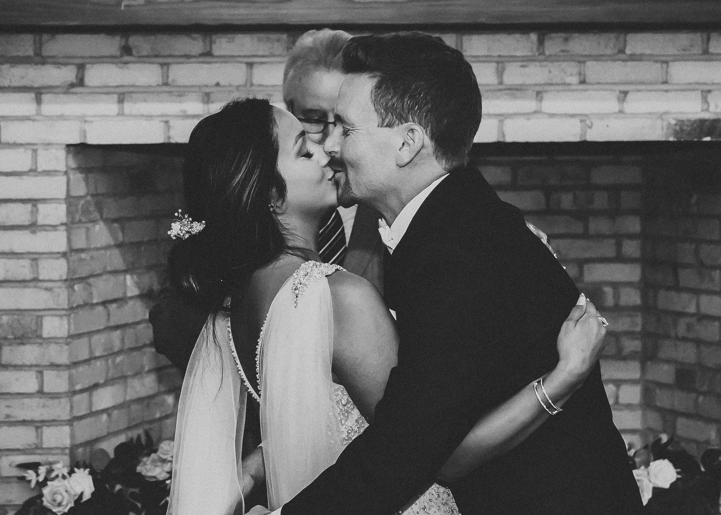 A black-and-white photo of a couple kissing during a wedding ceremony, with a brick fireplace and flowers in the background.