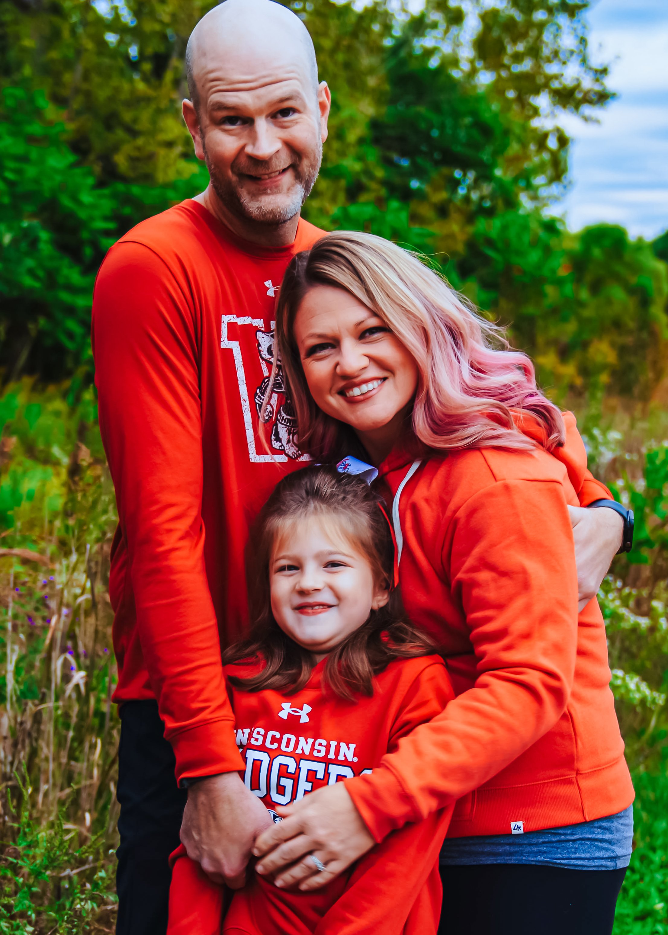 A smiling family of three wearing Wisconsin Badgers shirts outdoors with greenery and trees in the background.