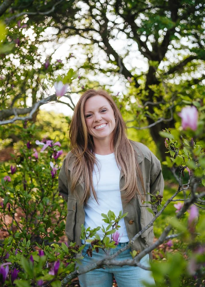 A smiling woman with long brown hair stands among green bushes and pink flowers outdoors on a sunny day.