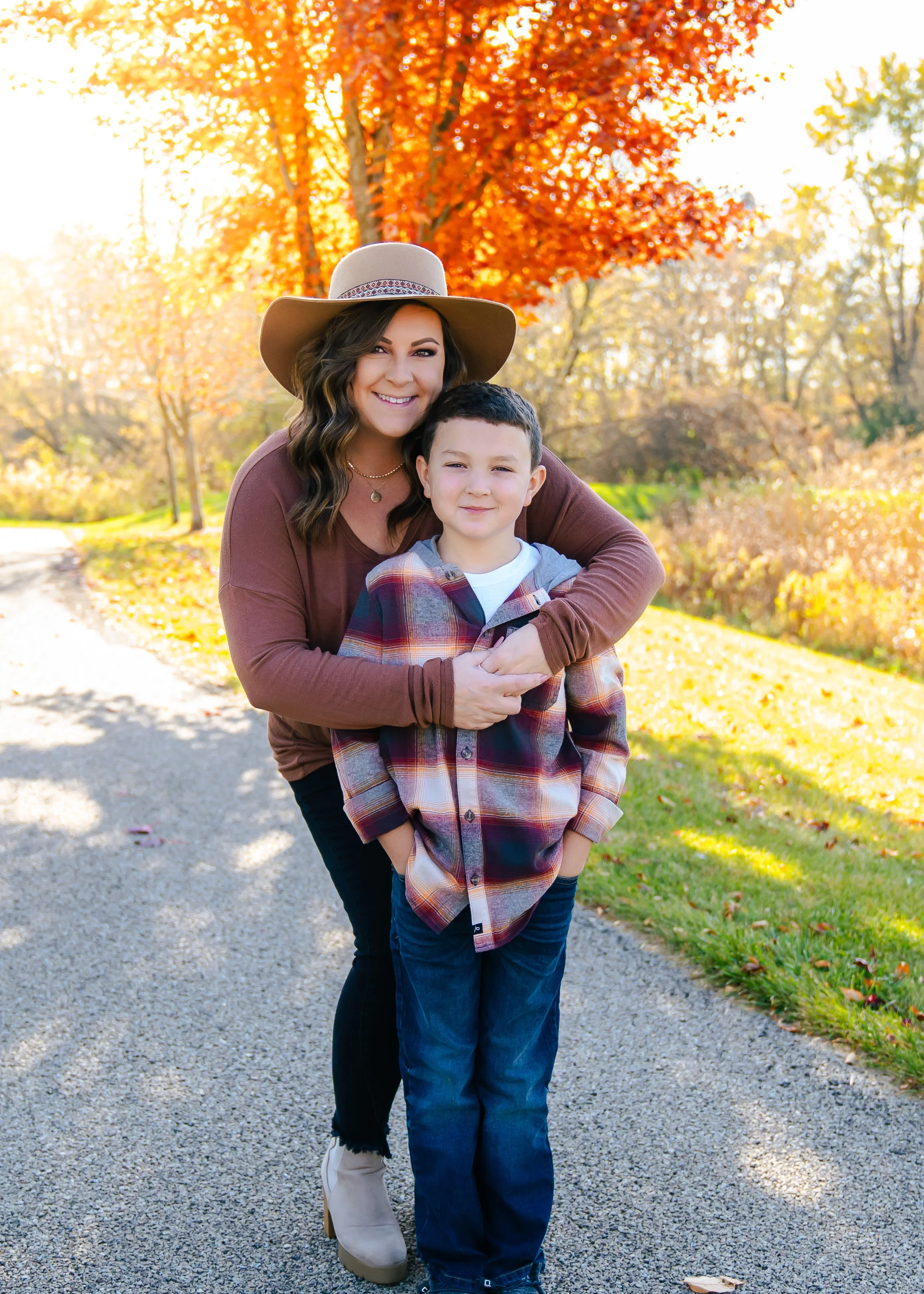 A woman and a boy are outdoors during fall, standing on a gravel path with autumn trees and colorful foliage in the background. The woman is wearing a wide-brimmed hat and a long-sleeve top, smiling and hugging the boy who is wearing a plaid shirt.