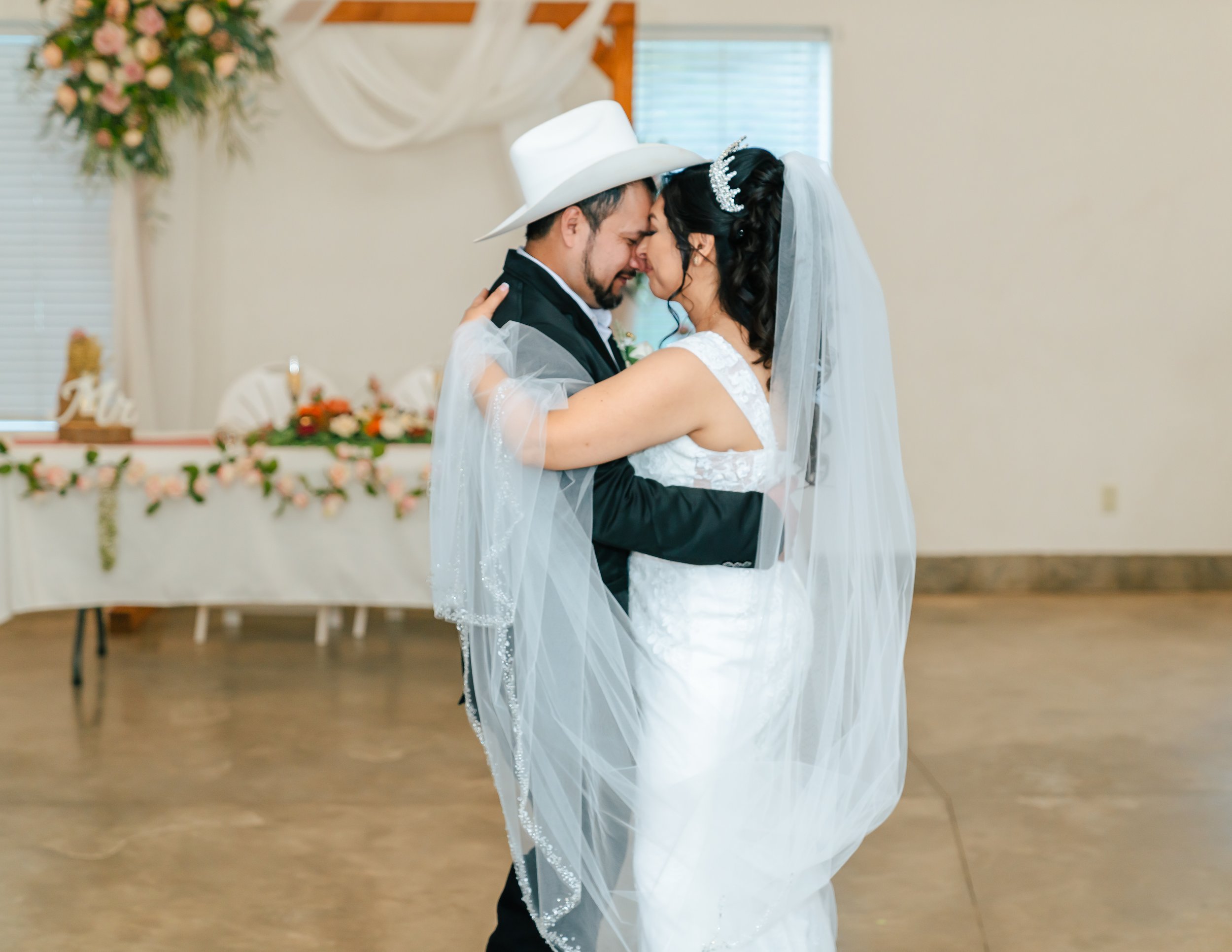 A bride and groom dancing closely on their wedding day, with the bride wearing a white wedding dress with a veil and tiara, and the groom wearing a black suit and white hat, in a decorated indoor venue with a table and floral decor in the background.
