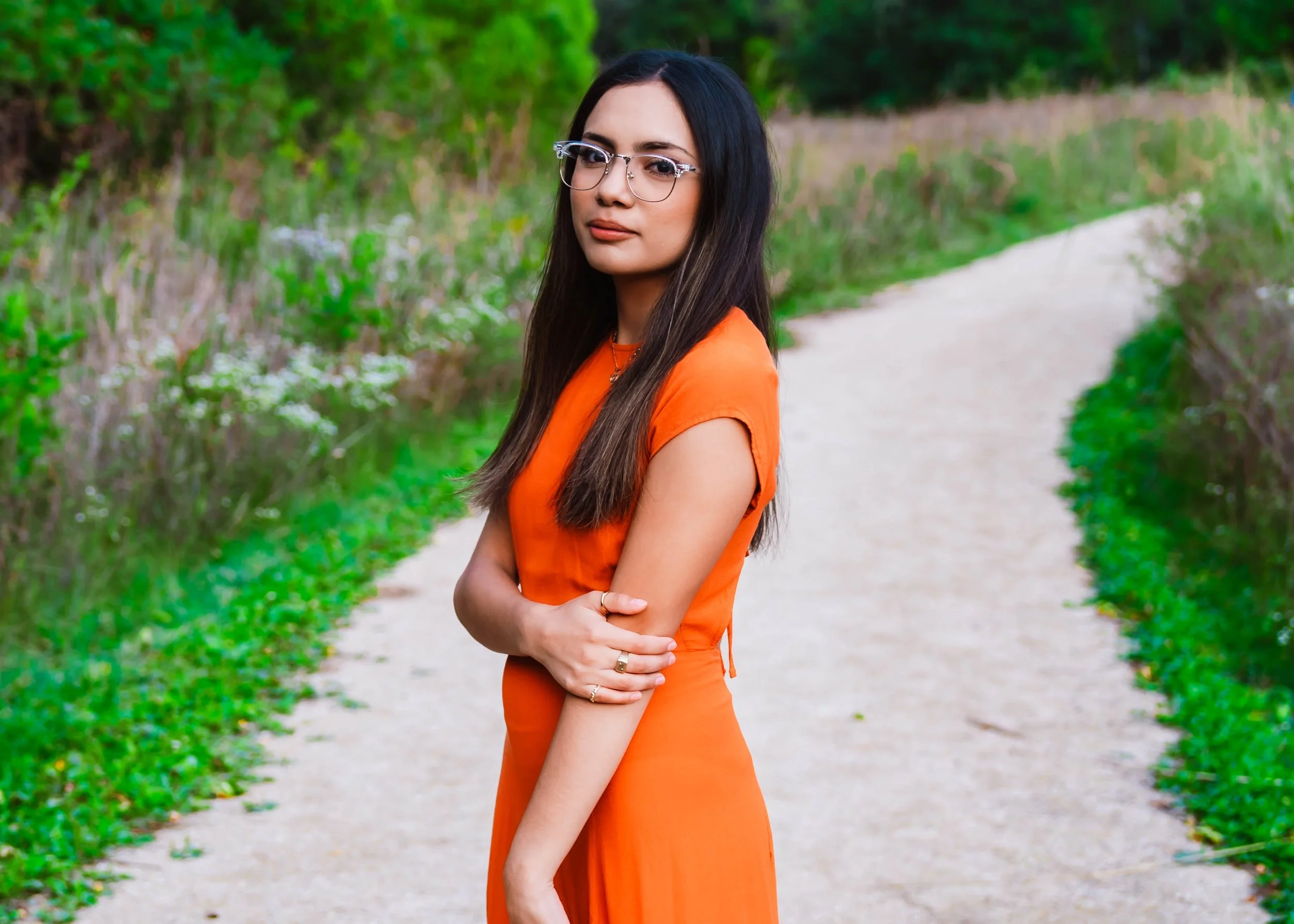 A woman with long dark hair and glasses wearing an orange dress standing on a winding dirt path surrounded by green foliage.