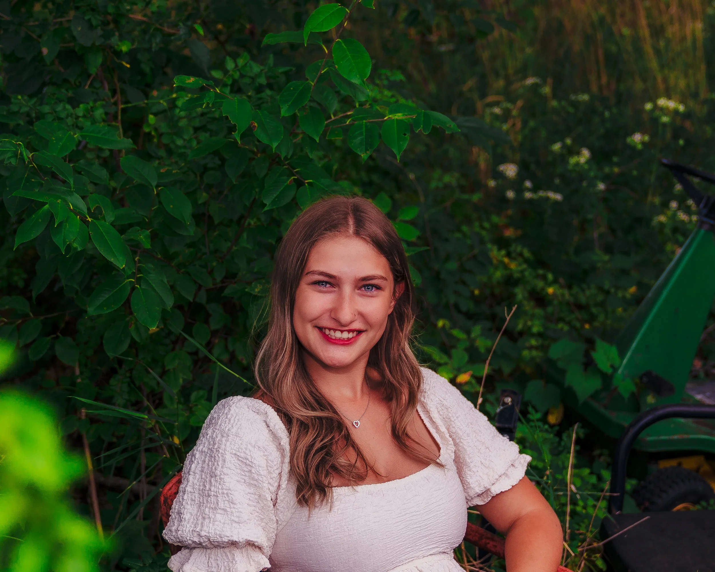 A young woman with long, wavy brown hair and blue eyes is smiling and sitting outdoors in front of green leafy bushes, wearing a white textured dress and a heart-shaped necklace.