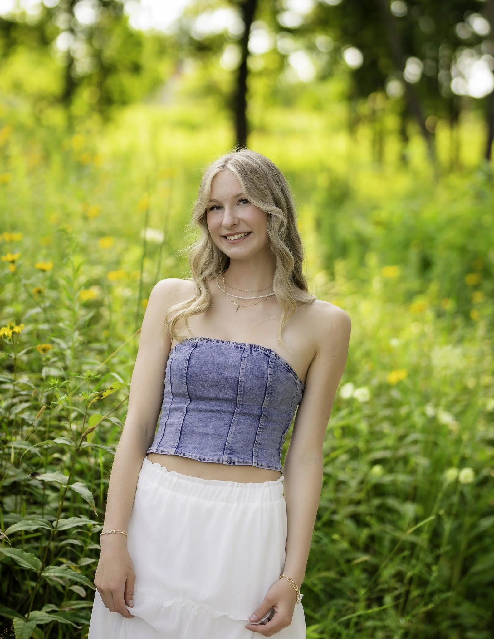 A young woman with blonde hair, smiling, outdoors in a green forest, wearing a strapless denim crop top and a white skirt.