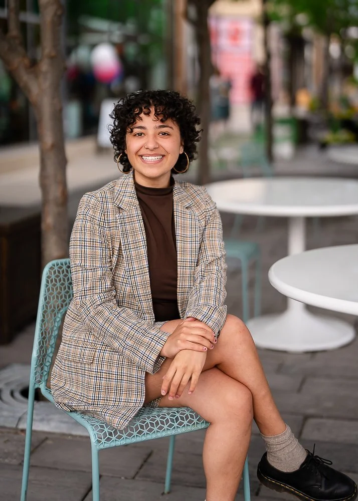 A young woman with curly hair smiling and sitting outside at a cafe on a light blue chair.