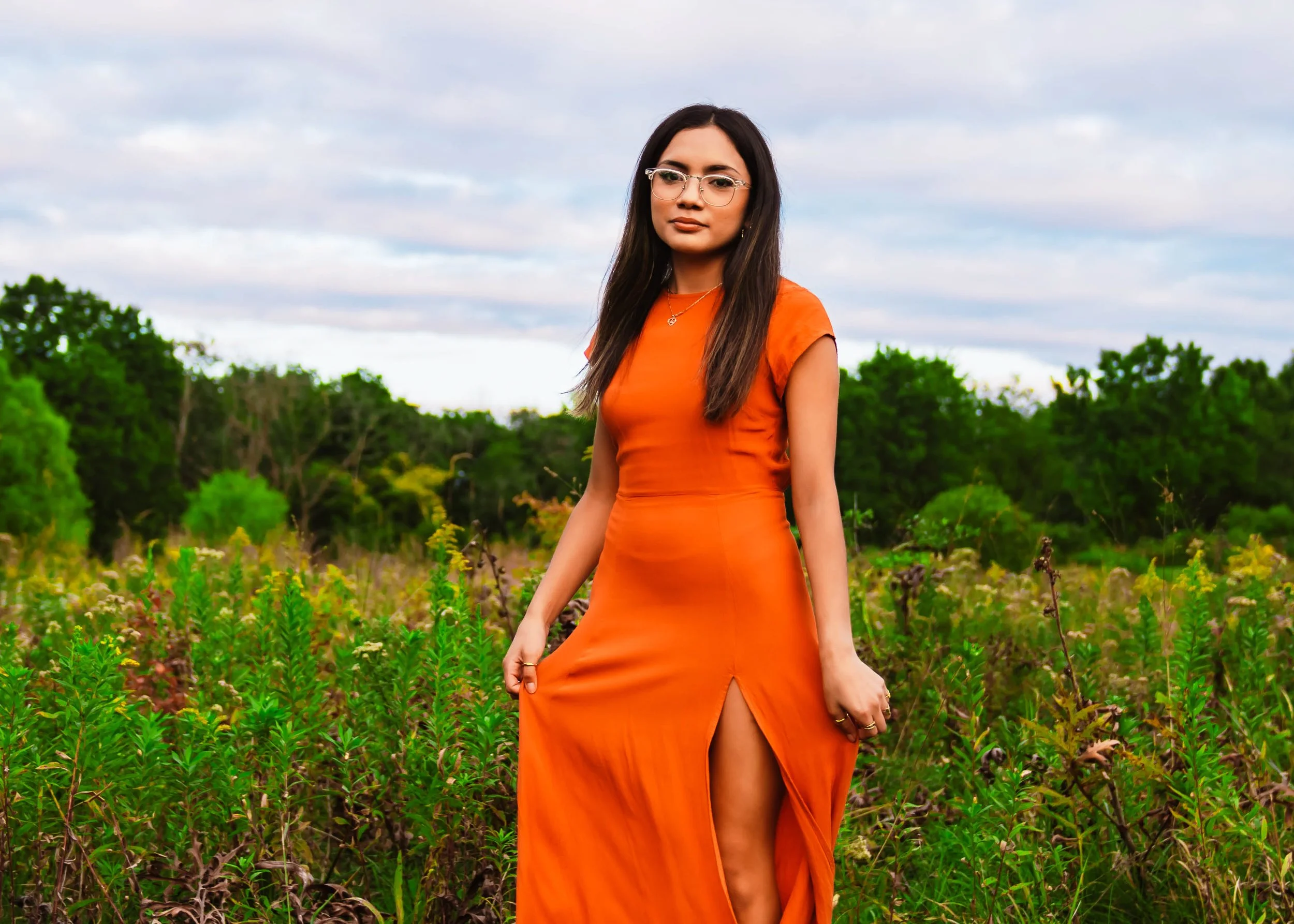 Woman in an orange dress standing in a grassy field with green trees and cloudy sky in the background.