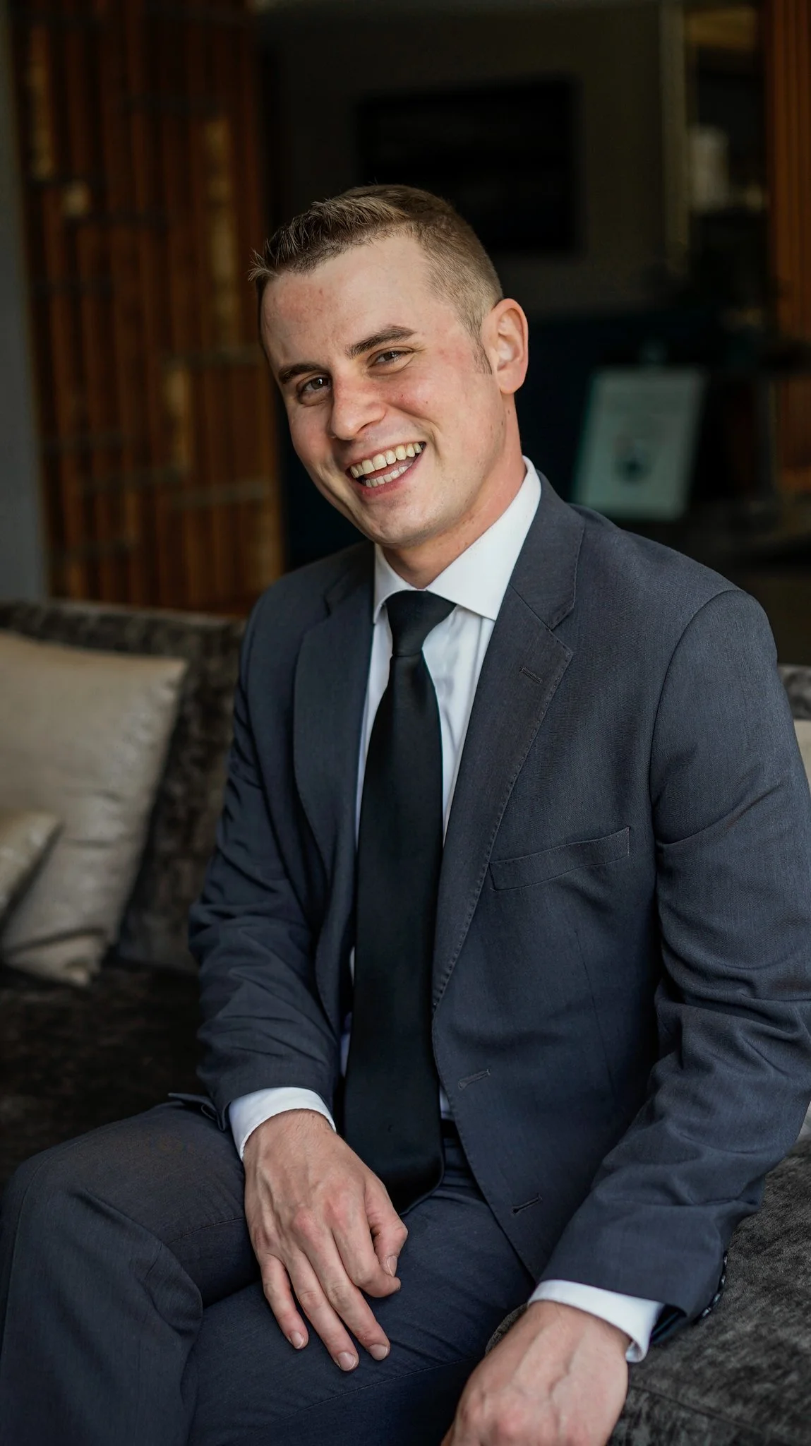 A man in a dark gray suit, white dress shirt, and black tie, smiling and sitting on a couch in a room with wood paneling and decorative pillows.