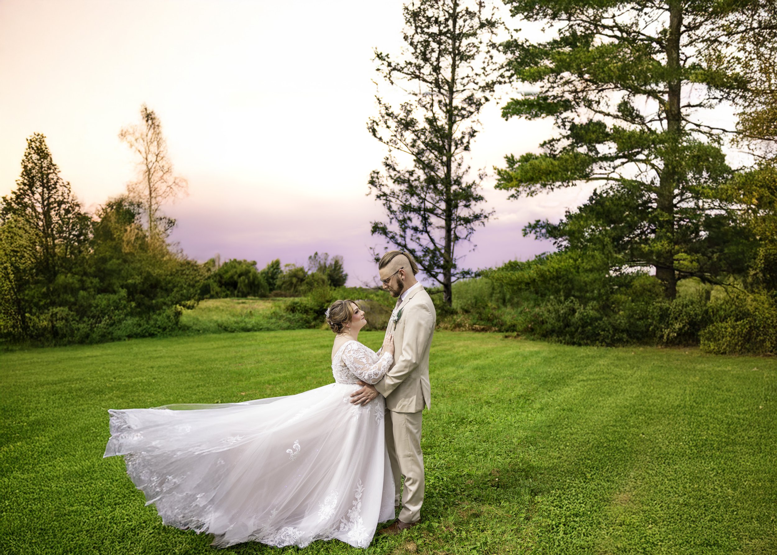 A bride and groom standing closely together on a green lawn with trees and a colorful sky in the background during sunset.